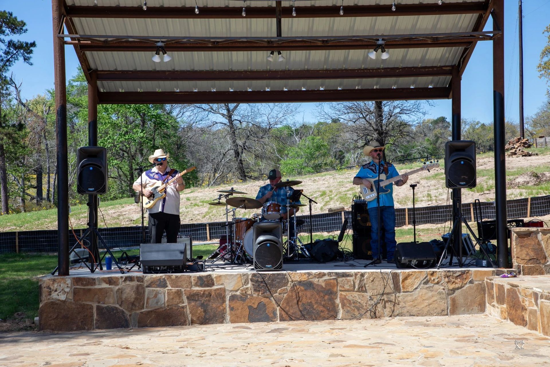 Three musicians performing on an outdoor stage under a metal canopy, with drums and speakers.