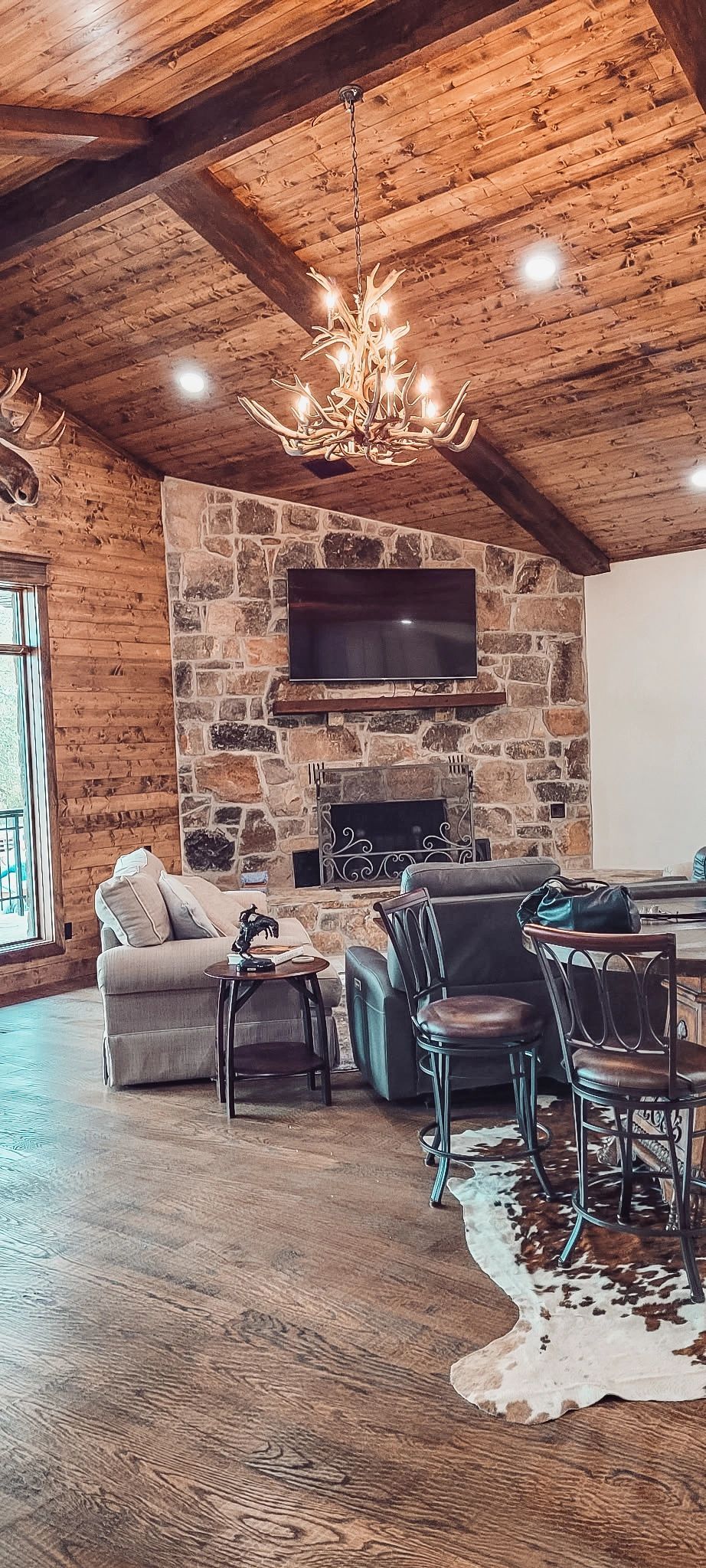 Rustic living room with stone fireplace, wall-mounted TV, chandelier, and wooden ceiling