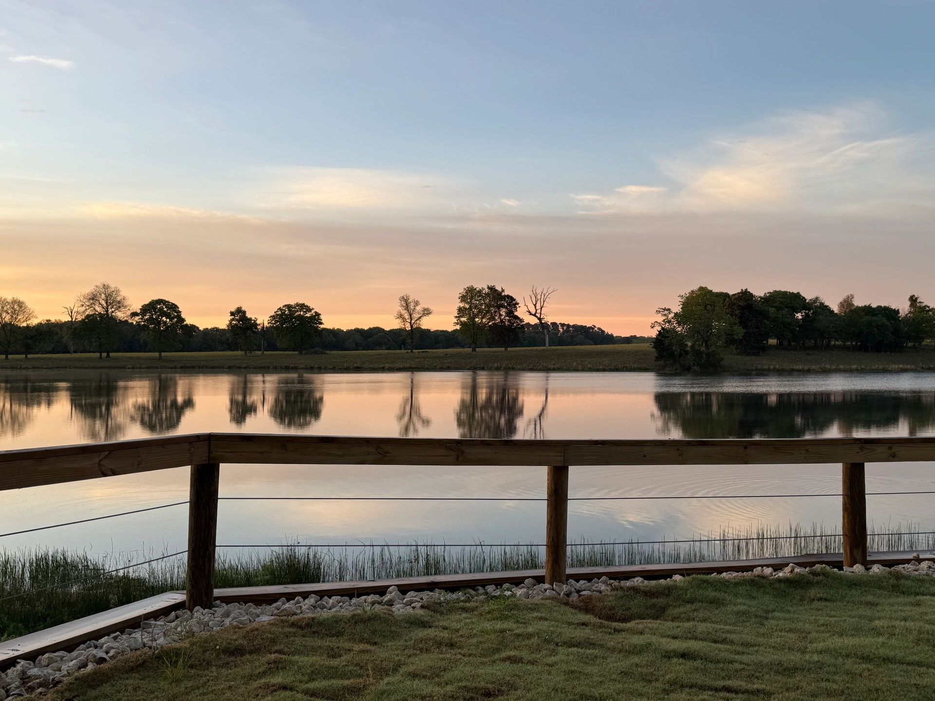 Sunset over a calm lake with tree reflections and a wooden railing in the foreground