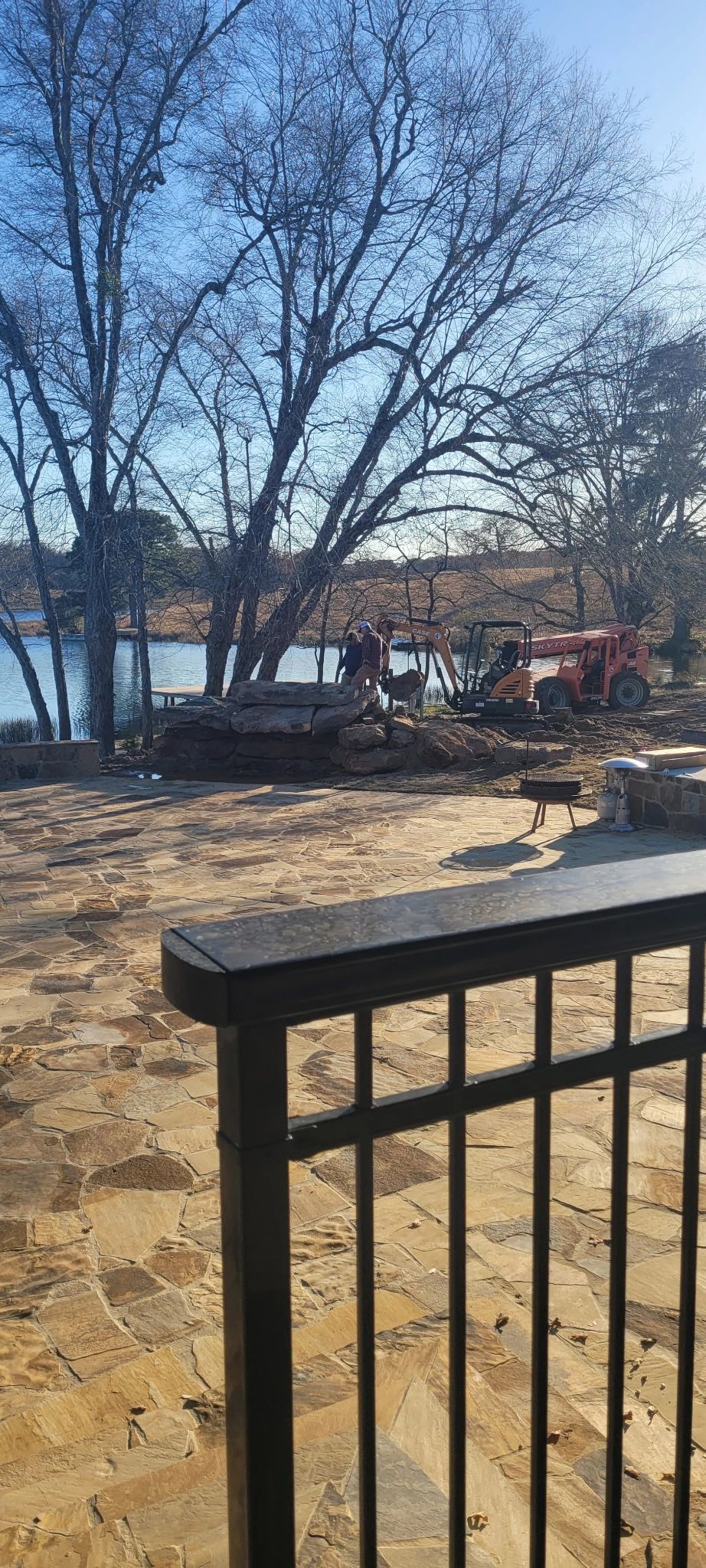 Sandy beach with leafless trees, parked cars, and a black railing in the foreground by the water.