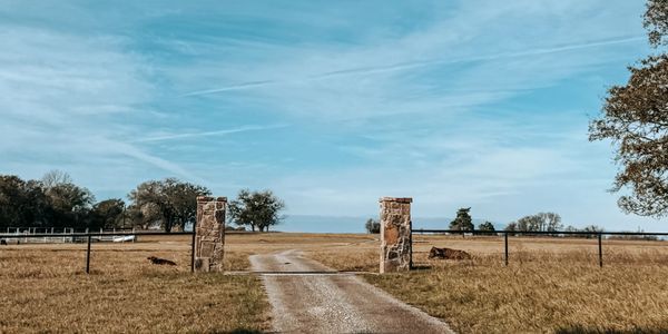 Rural dirt road leading through an open gate between stone pillars under a blue sky