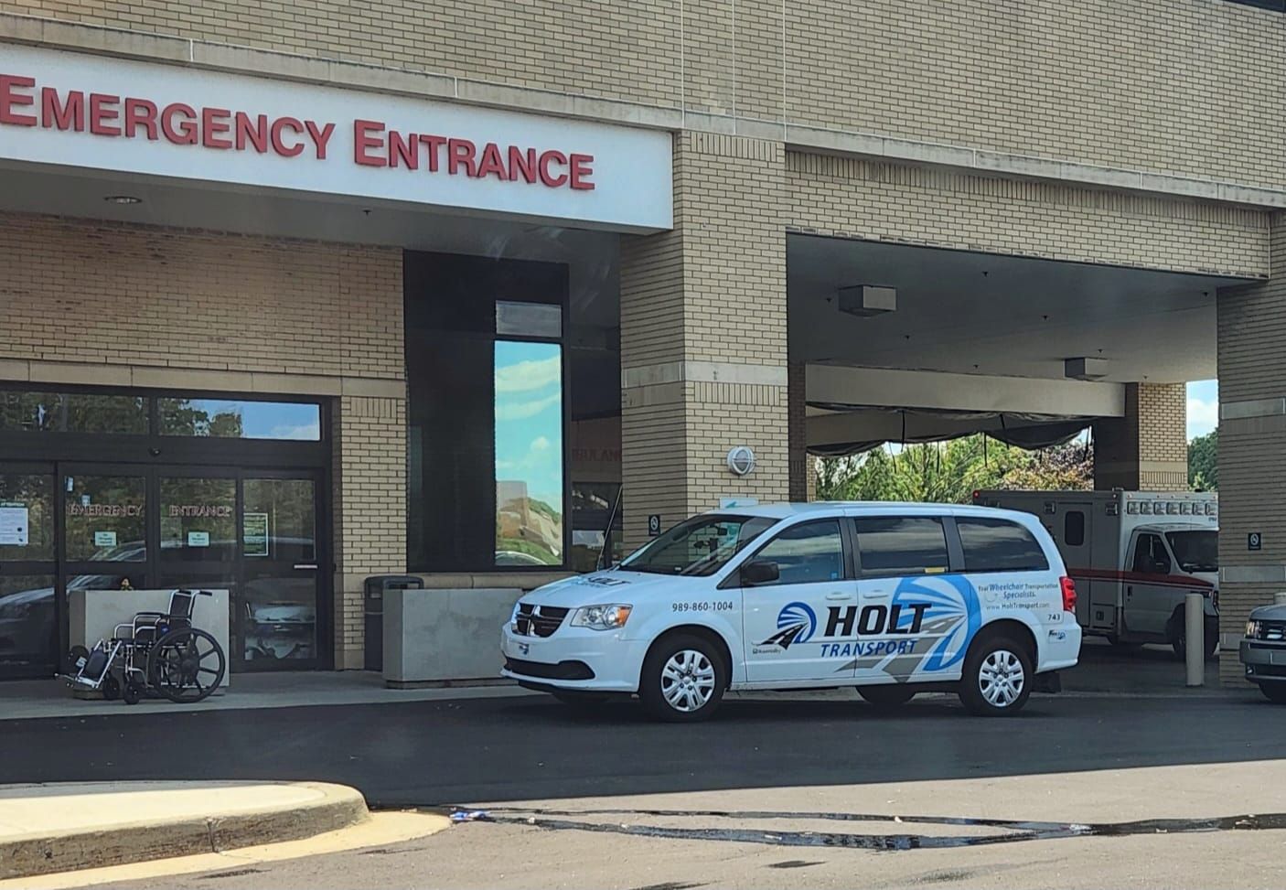 A white medical transport van parked outside a brick building under an Emergency Entrance sign, with a wheelchair nearby.