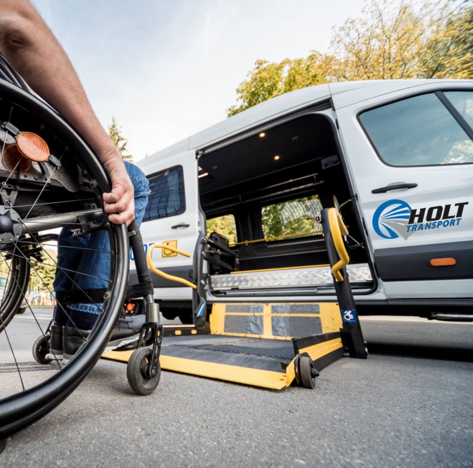 A person in a wheelchair prepares to use the lowered yellow lift of a white Holt Transport van.