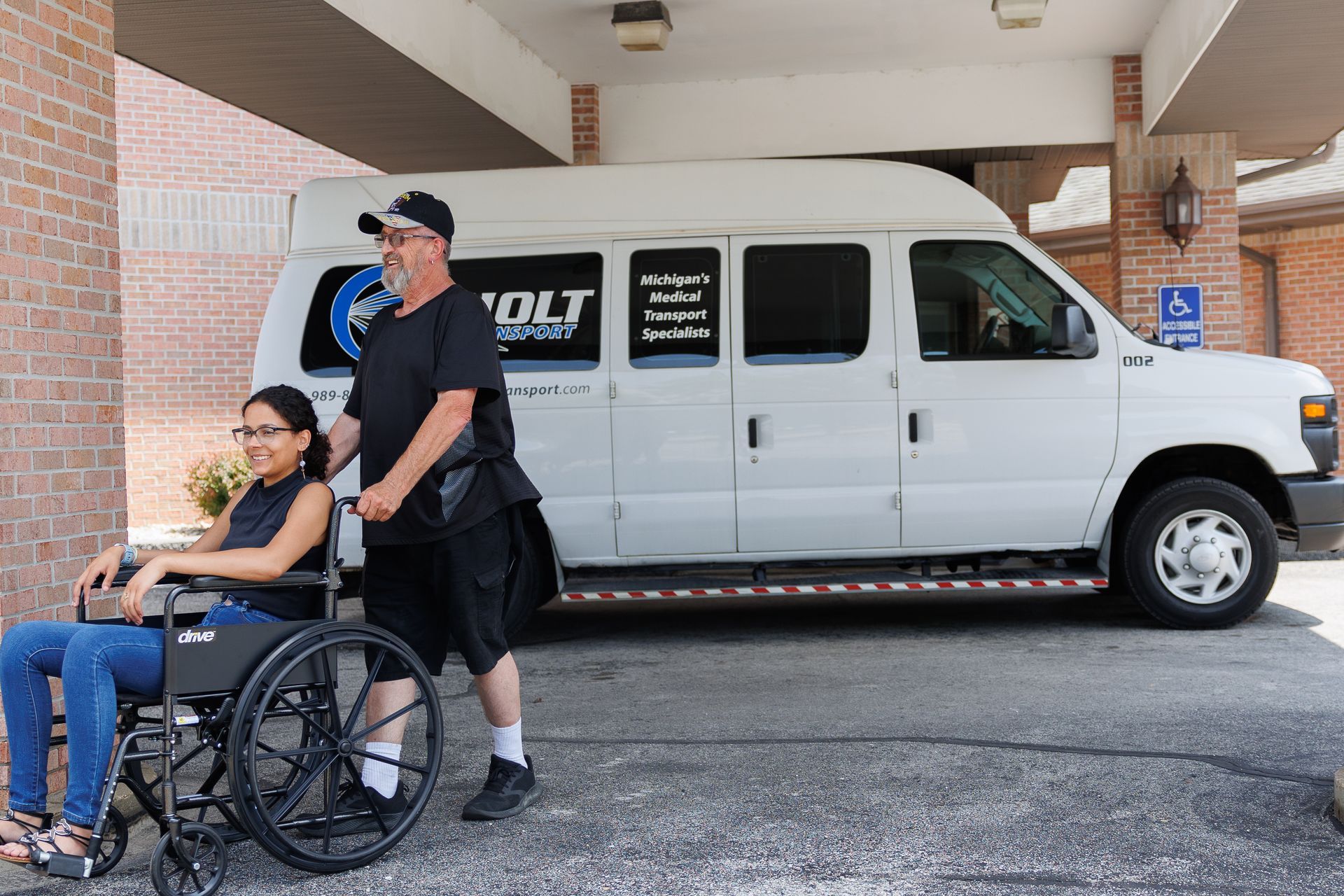 Person in wheelchair with flowers beside a uniformed man near a white van under a covered walkway