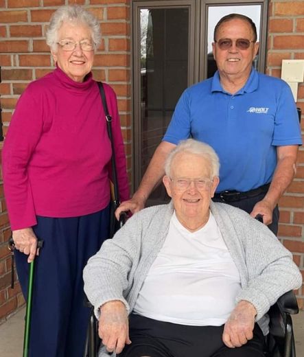Three adults on a porch, one seated in a wheelchair, smiling at the camera.