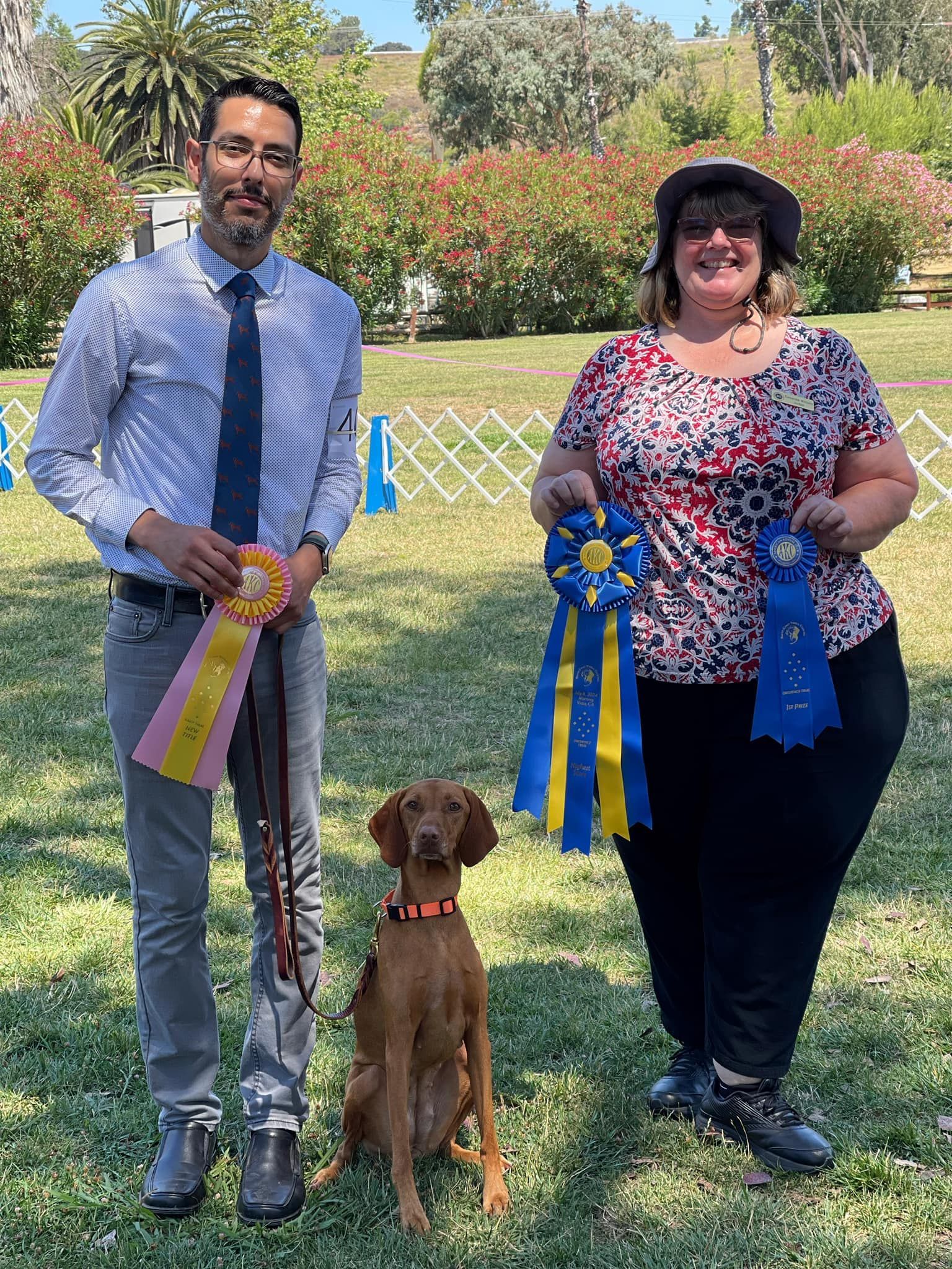 A man and a woman are standing next to a dog holding ribbons.