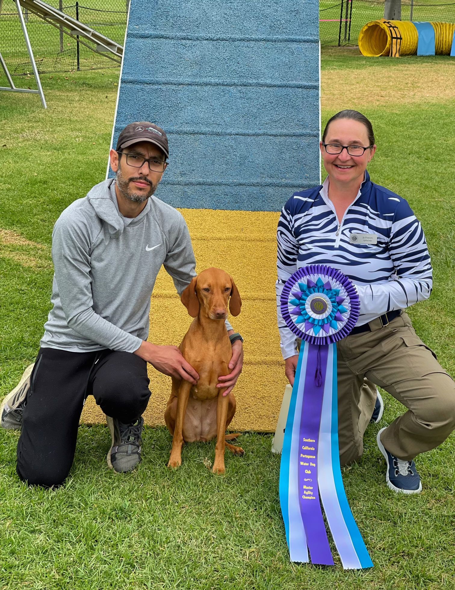A man and a woman are kneeling next to a dog holding a ribbon.