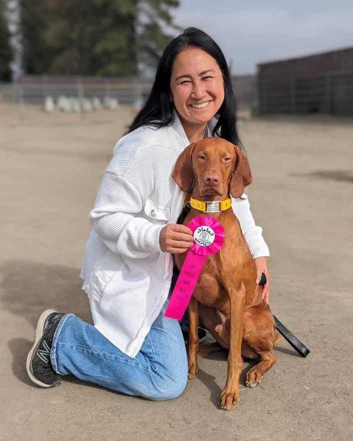 A woman is kneeling down next to a brown dog holding a ribbon.