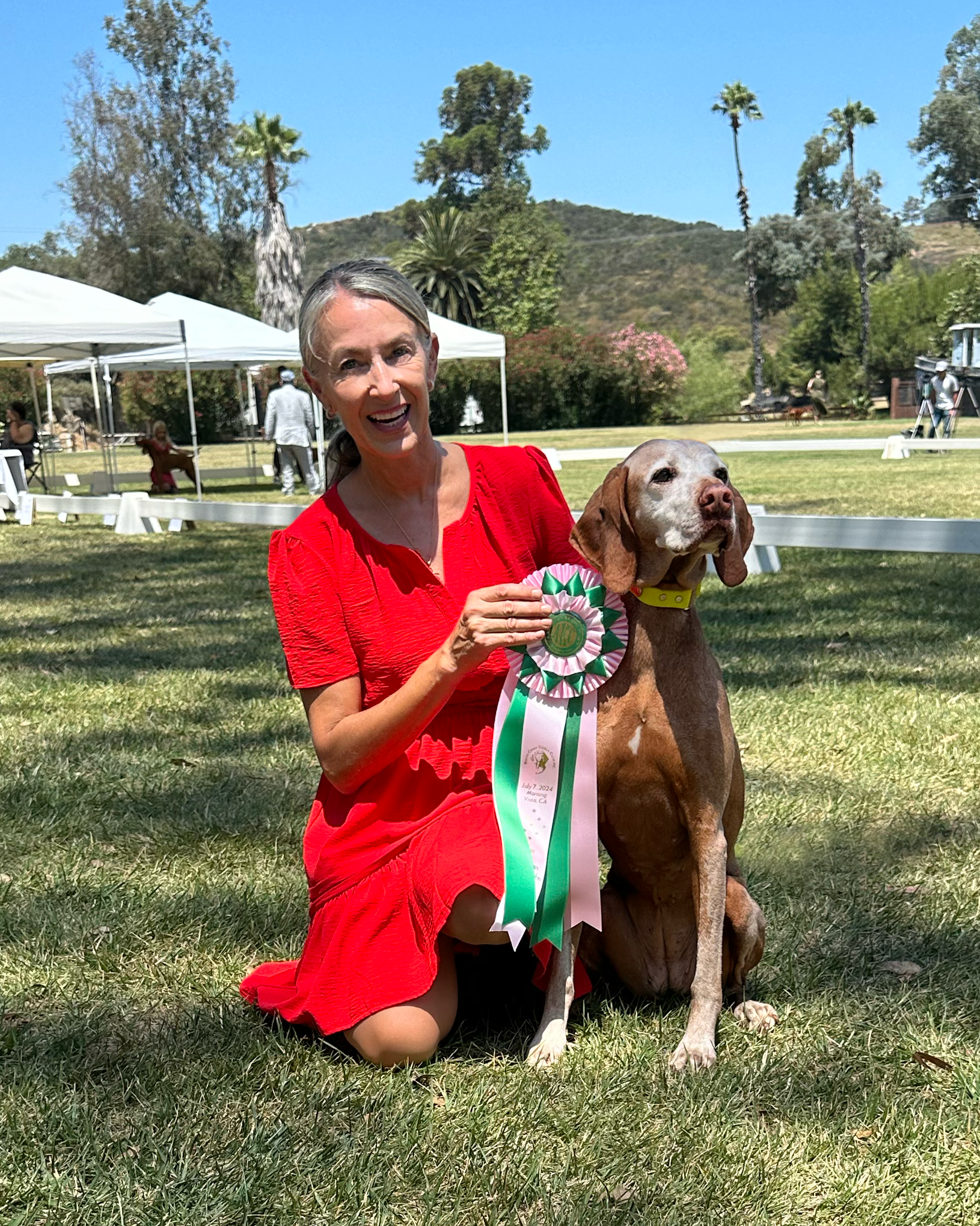 A woman in a red dress is kneeling next to a brown dog holding a ribbon.