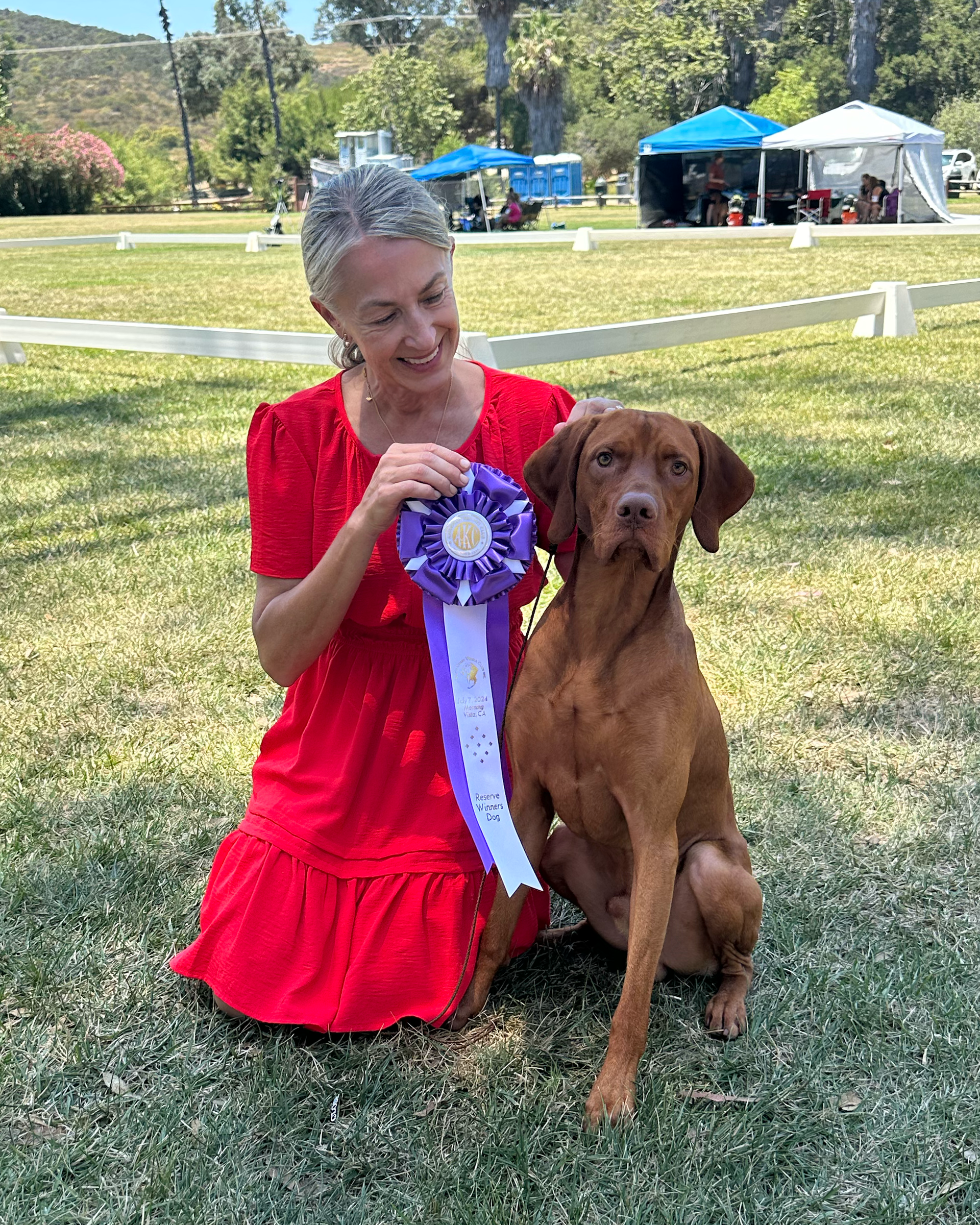 A woman is kneeling down next to a dog holding a ribbon.