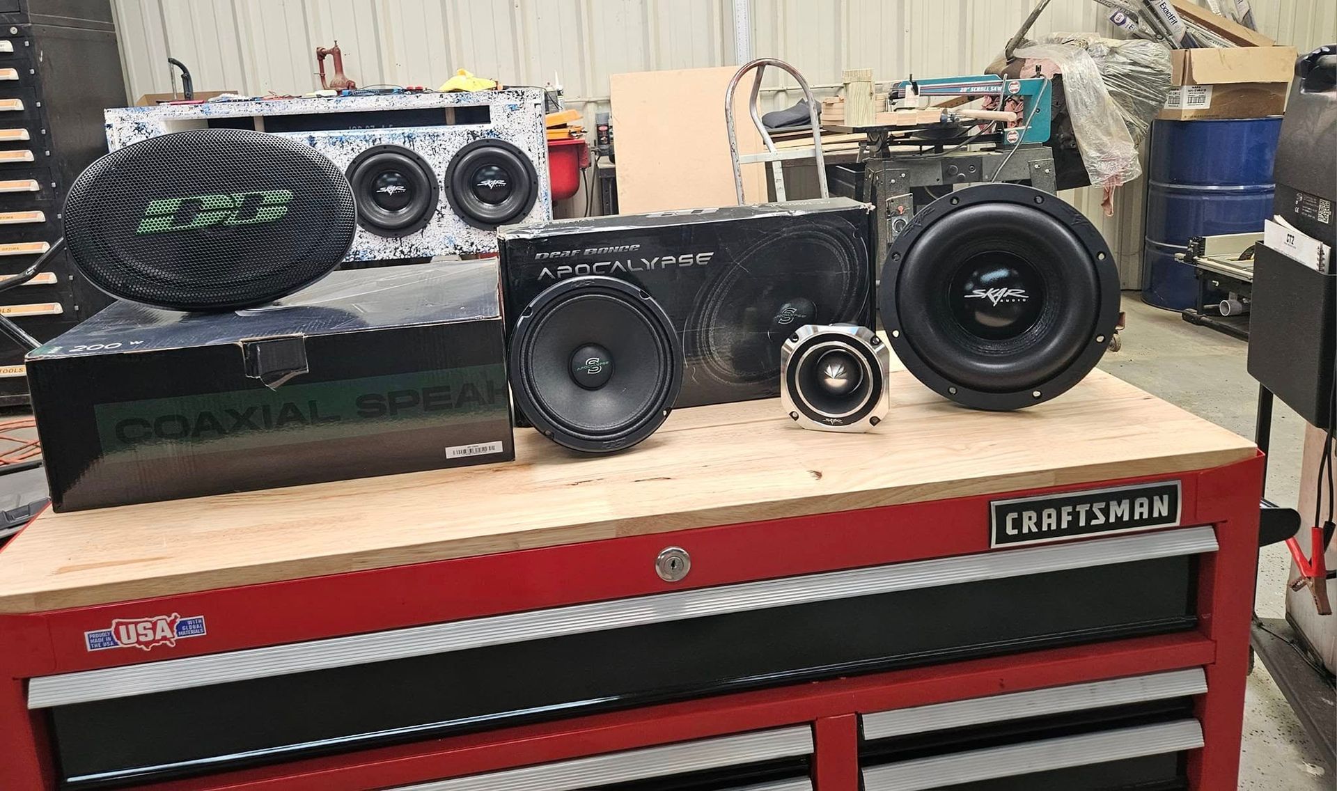 Car audio speakers and subwoofers arranged on a red Craftsman tool chest in a workshop.