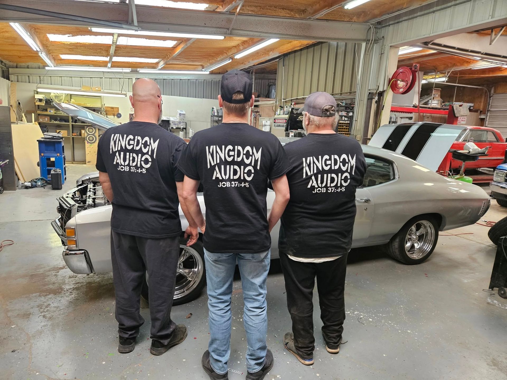 Three people in black shirts stand in a garage beside a silver classic car and workbench tools.