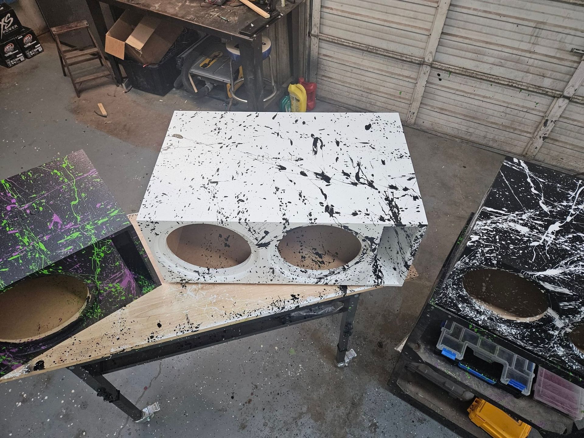 Three rectangular speaker enclosures in a workshop, with a white box in the center and two black-speckled boxes on either side.
