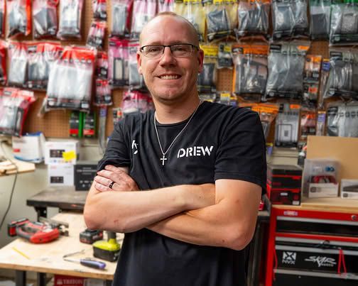 Smiling man in a black shirt stands in a tool shop with shelves of packaged items behind him