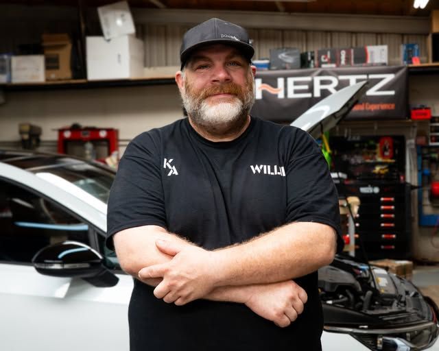 Mechanic in black shirt and cap standing in auto shop beside a car with its hood open