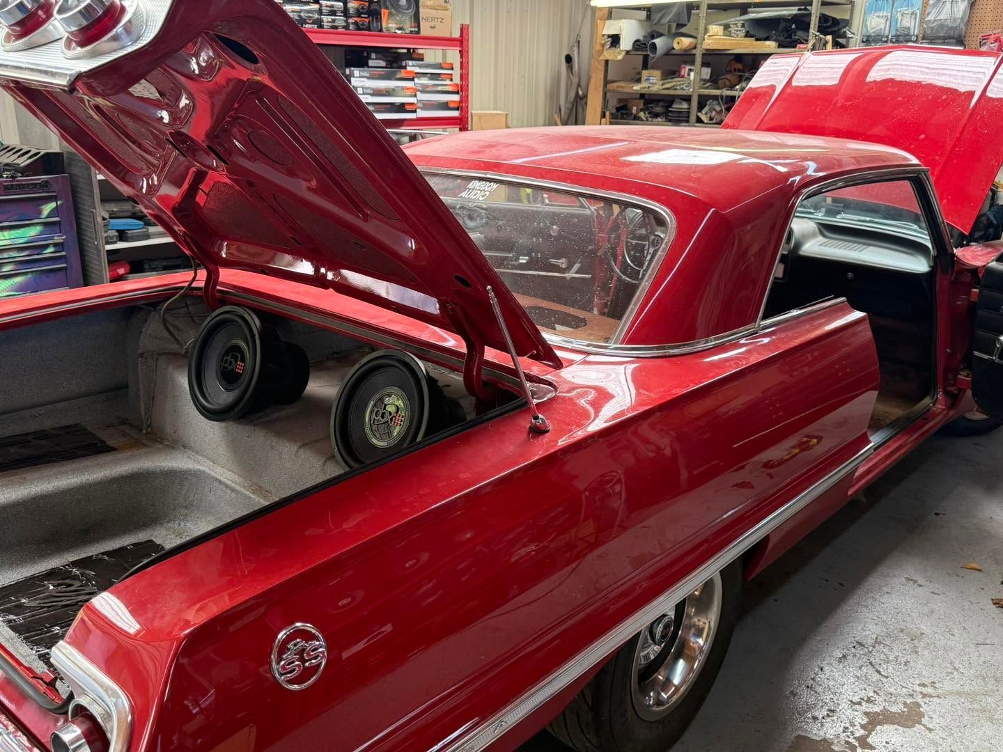 Red classic car in a garage with its trunk open, showing large speaker boxes inside and the door open.