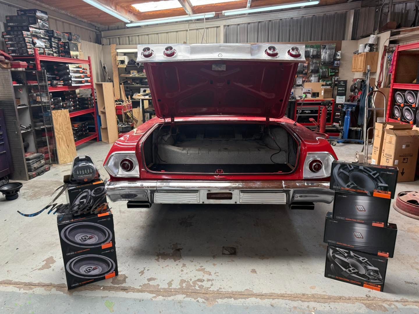 Red classic car with trunk open in a workshop, flanked by stacked speakers on both sides.