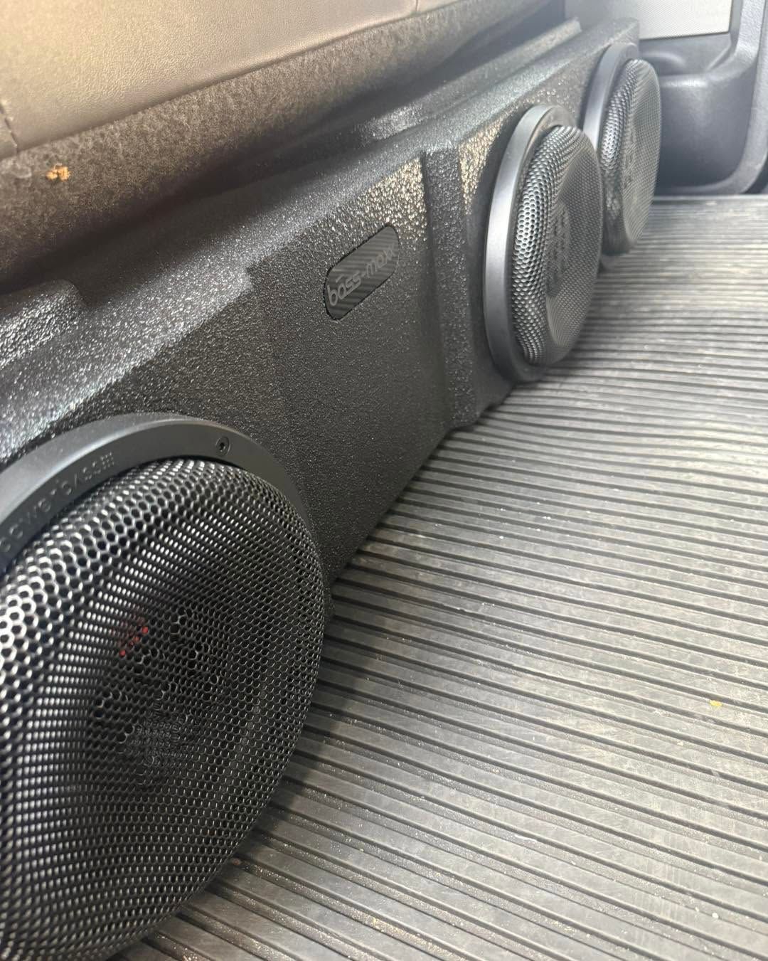Three black subwoofer speakers mounted in a vehicle cargo area on a ribbed floor mat