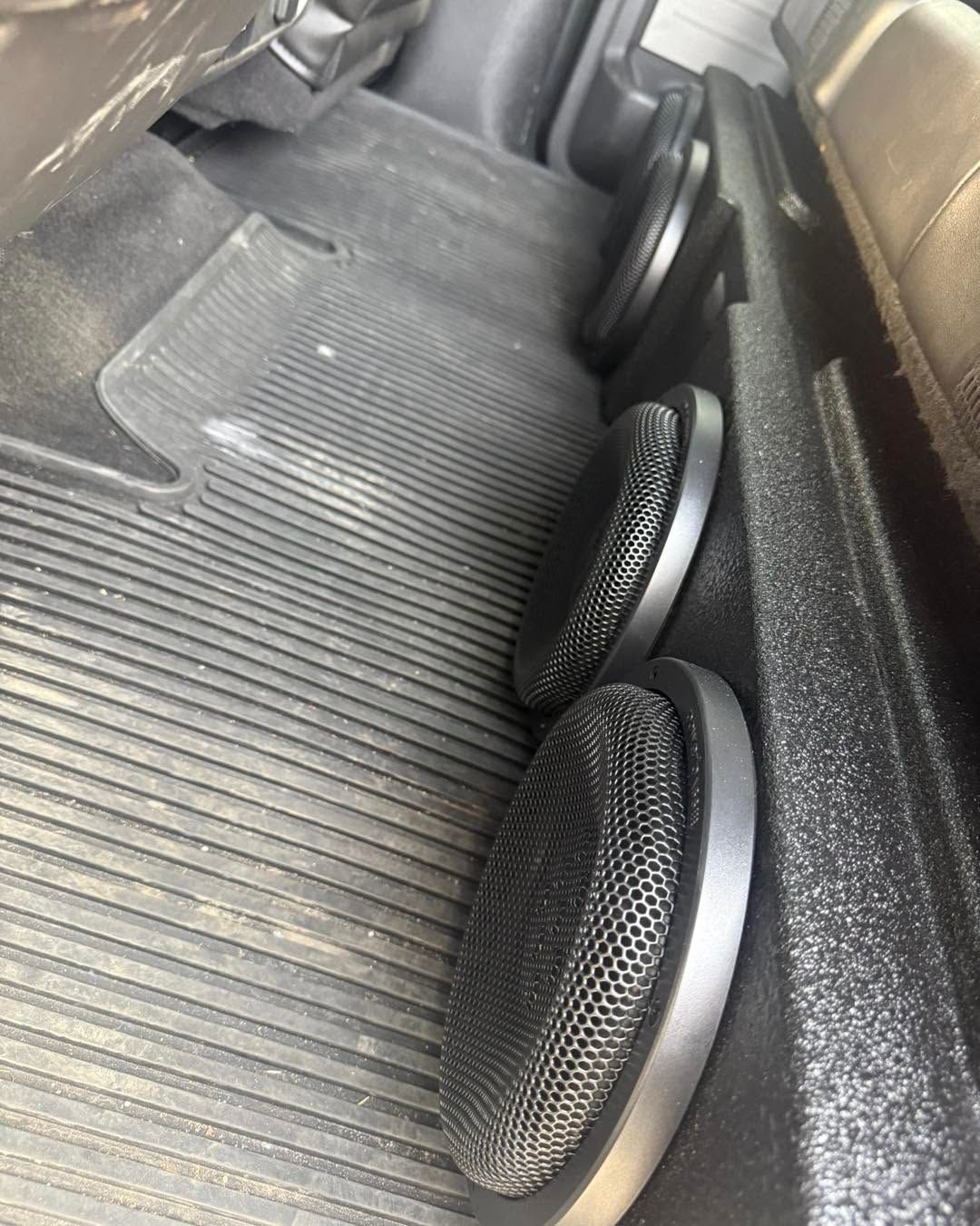 Two black round speakers mounted in the backseat of a vehicle with a ribbed gray floor mat