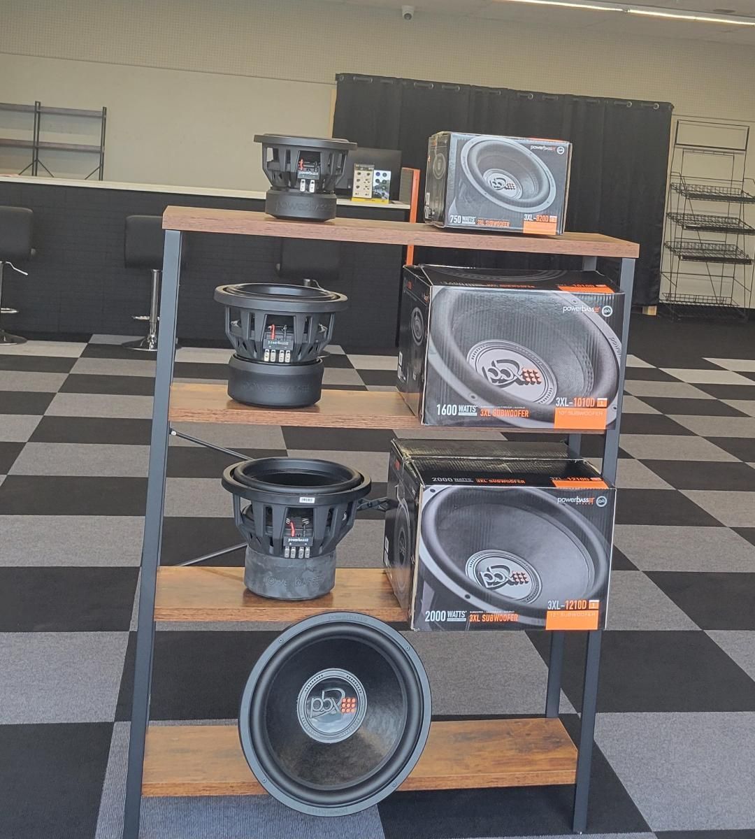 Car audio subwoofers displayed on a wooden shelf in a showroom with a checkered floor.