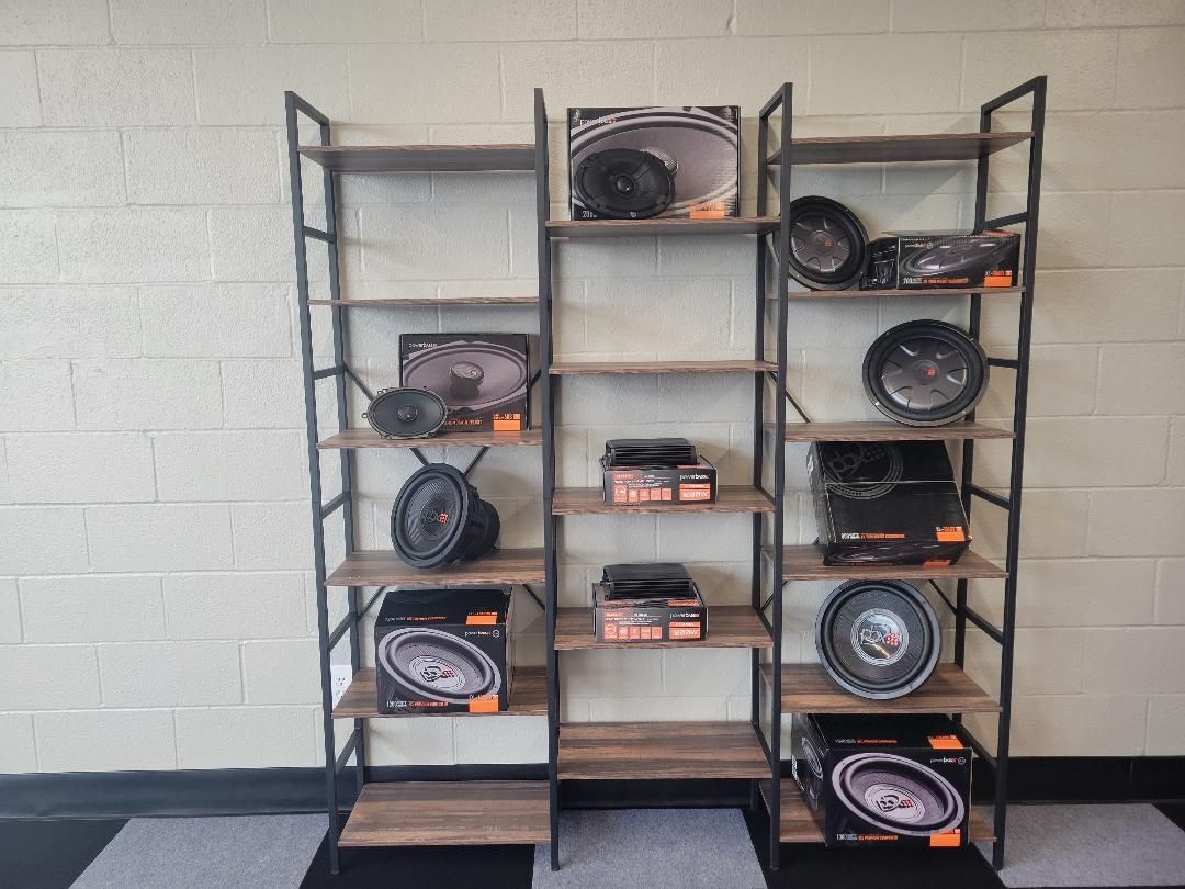 Black metal shelving with boxed car audio speakers and subwoofers against a gray cinderblock wall.