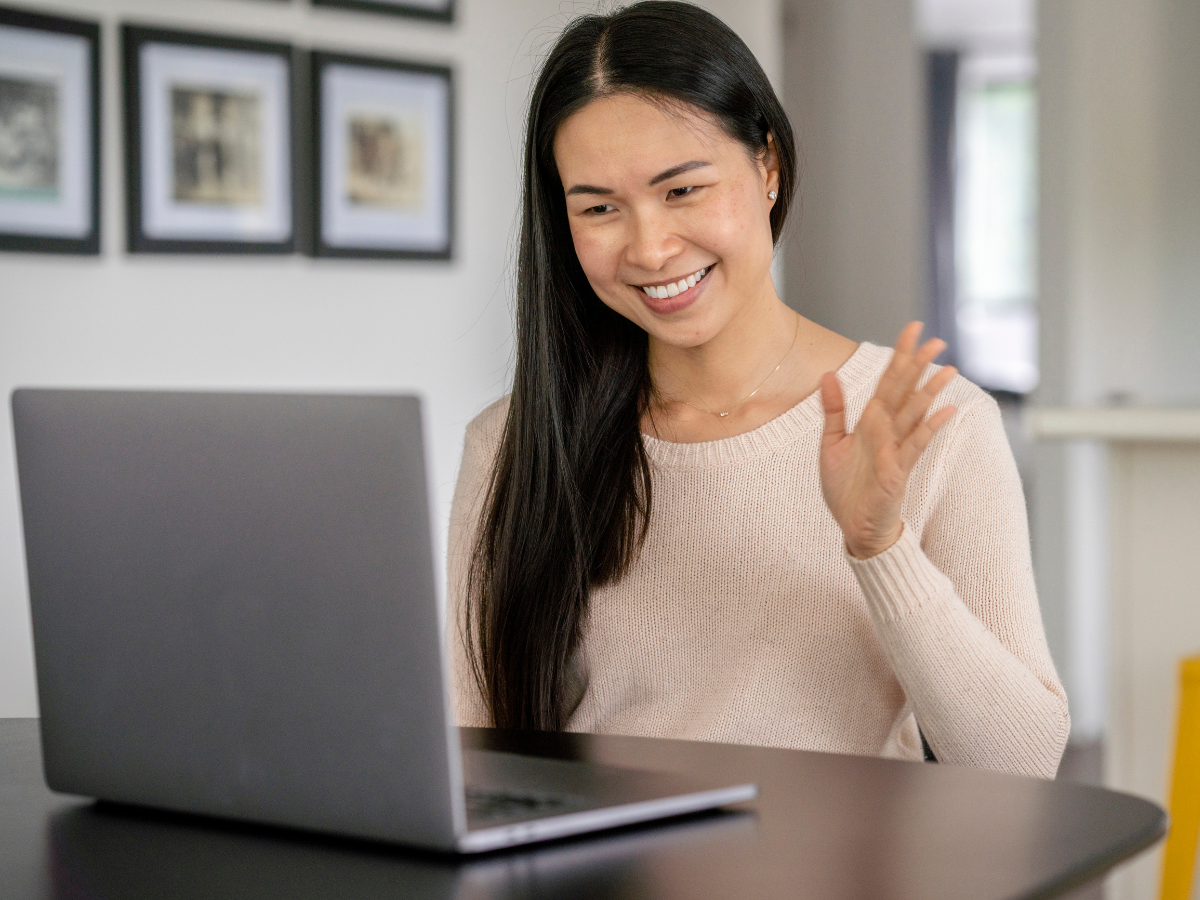 A person in a beige sweater sits at a table, smiling and waving at a laptop screen during a video call.