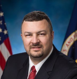 A man in a suit and red patterned tie poses for an official portrait with US flags in the background.
