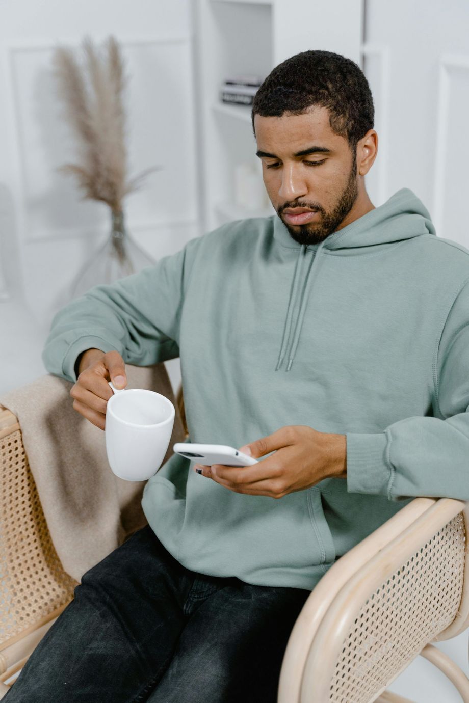 A person wearing a light green hoodie sits in a rattan chair, holding a white mug while looking at a smartphone.