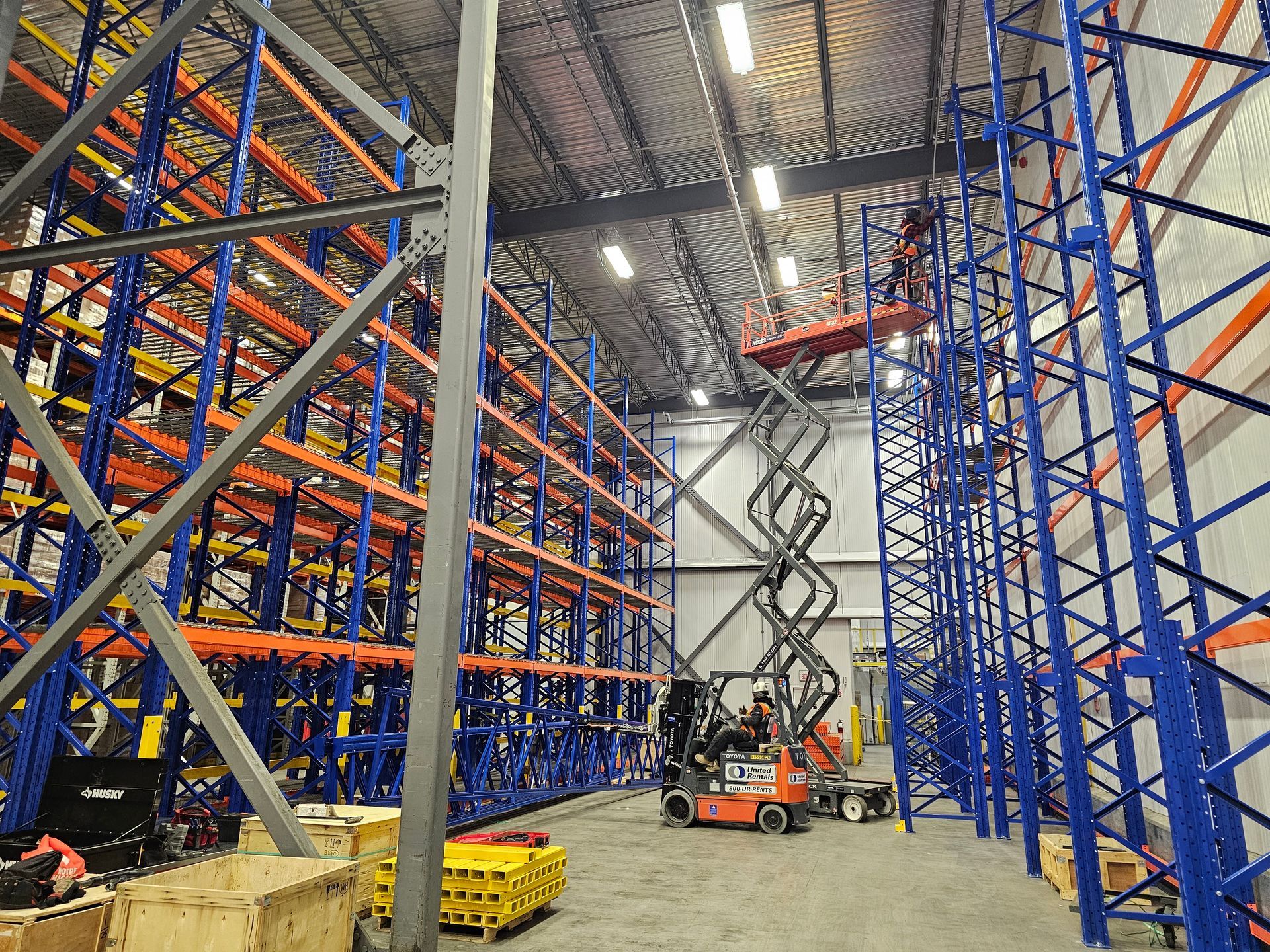 Services d'installation de rayonnages à l'intérieur d'un entrepôt équipé de rayonnages hauts bleus et orange, avec des ouvriers travaillant sur une plate-forme élévatrice.
