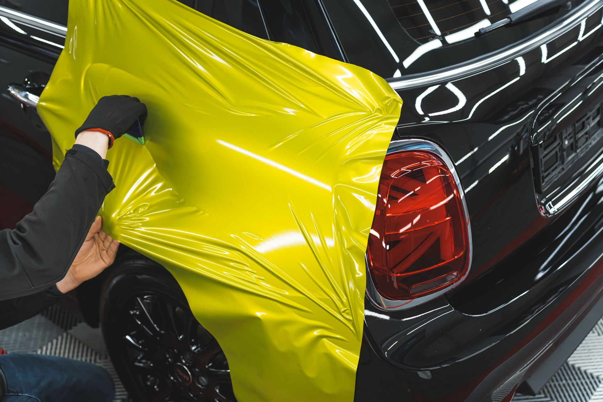 Person applying yellow vinyl wrap to the rear of a black car, near the taillight.