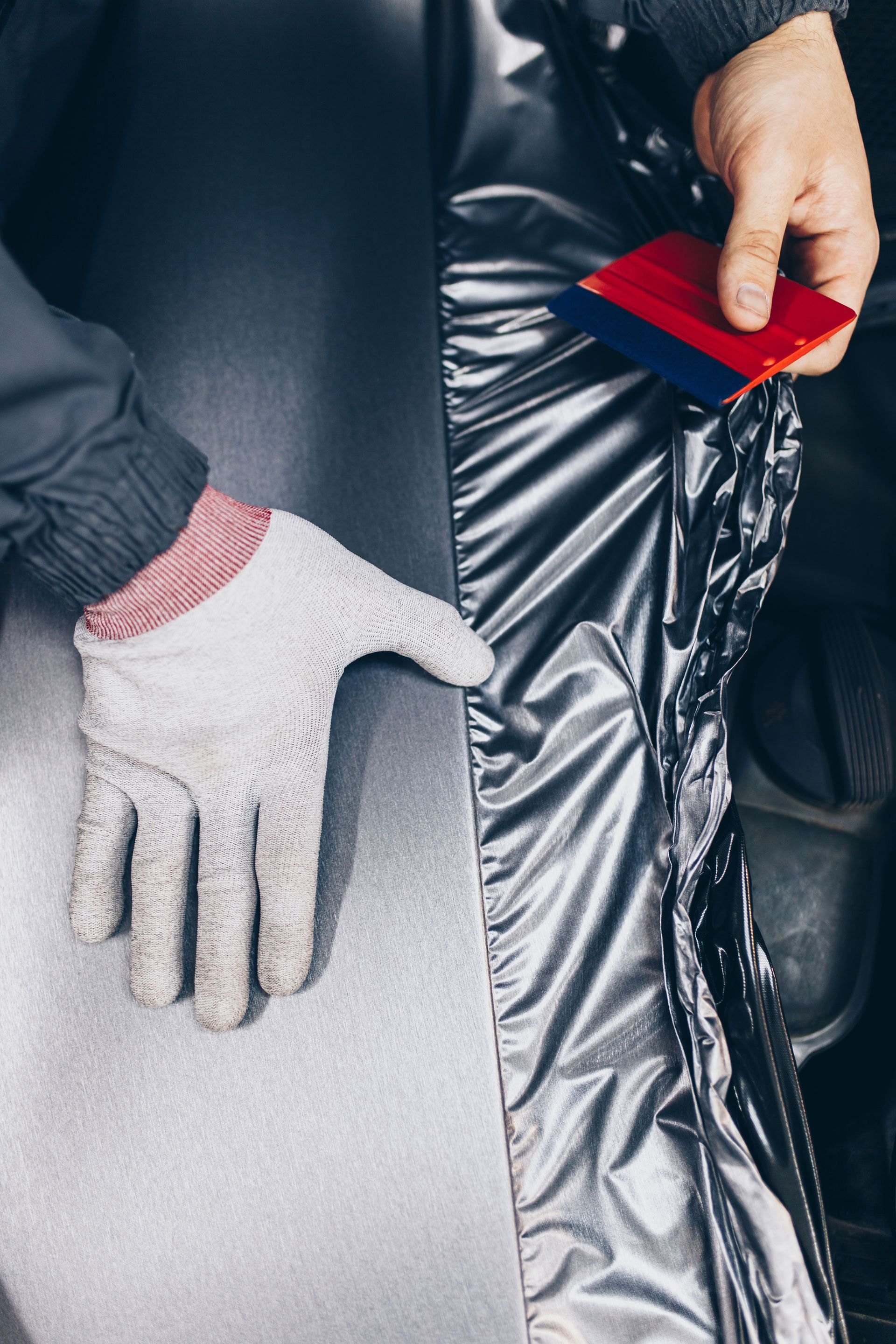 Person smoothing silver film onto a surface with a red squeegee, wearing work gloves.