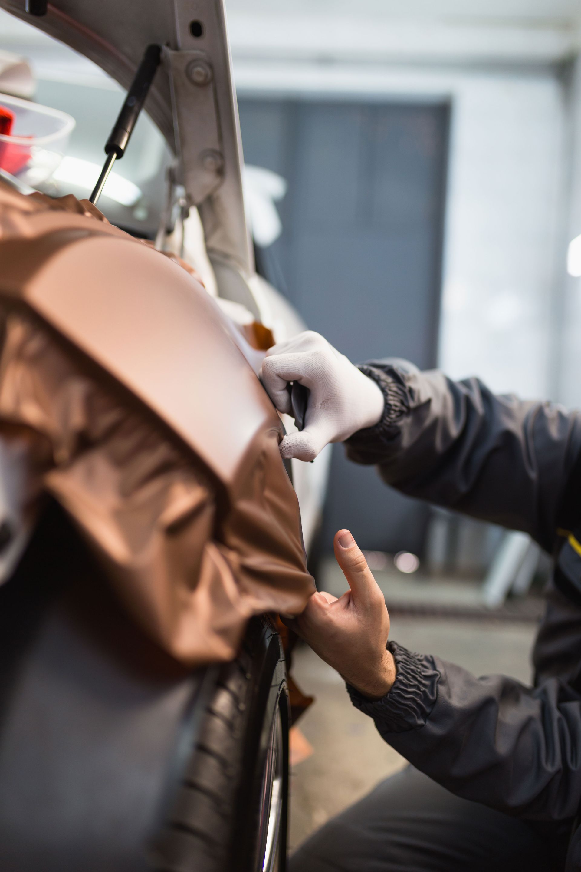 Person in gloves applying brown vinyl wrap to a car fender in a garage.