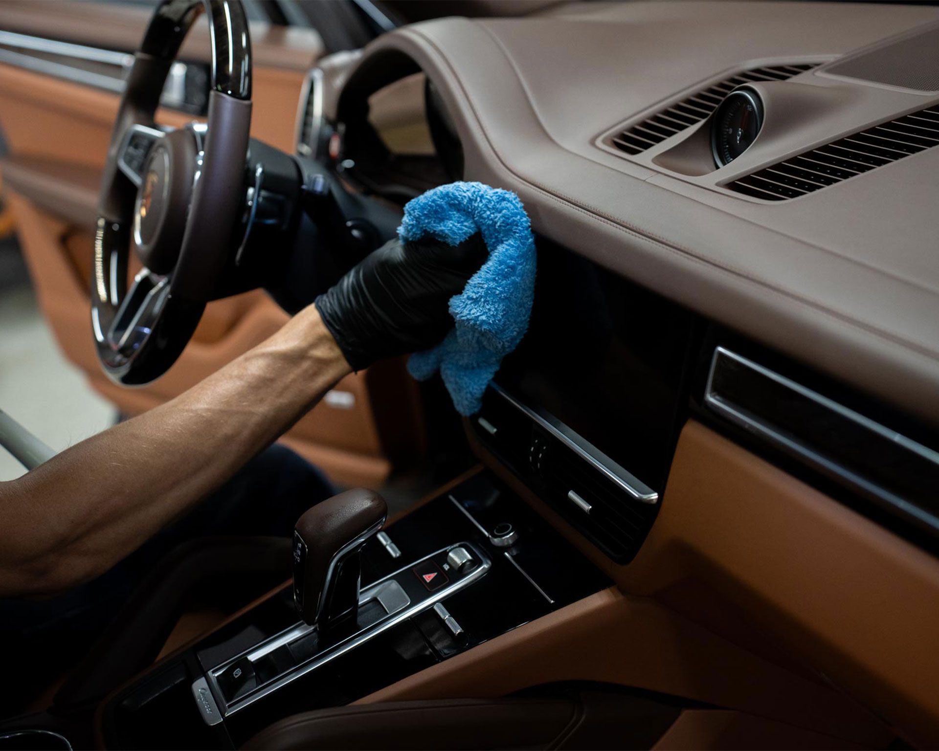 Gloved hand wiping a car dashboard with a blue cloth; interior view with brown leather details.