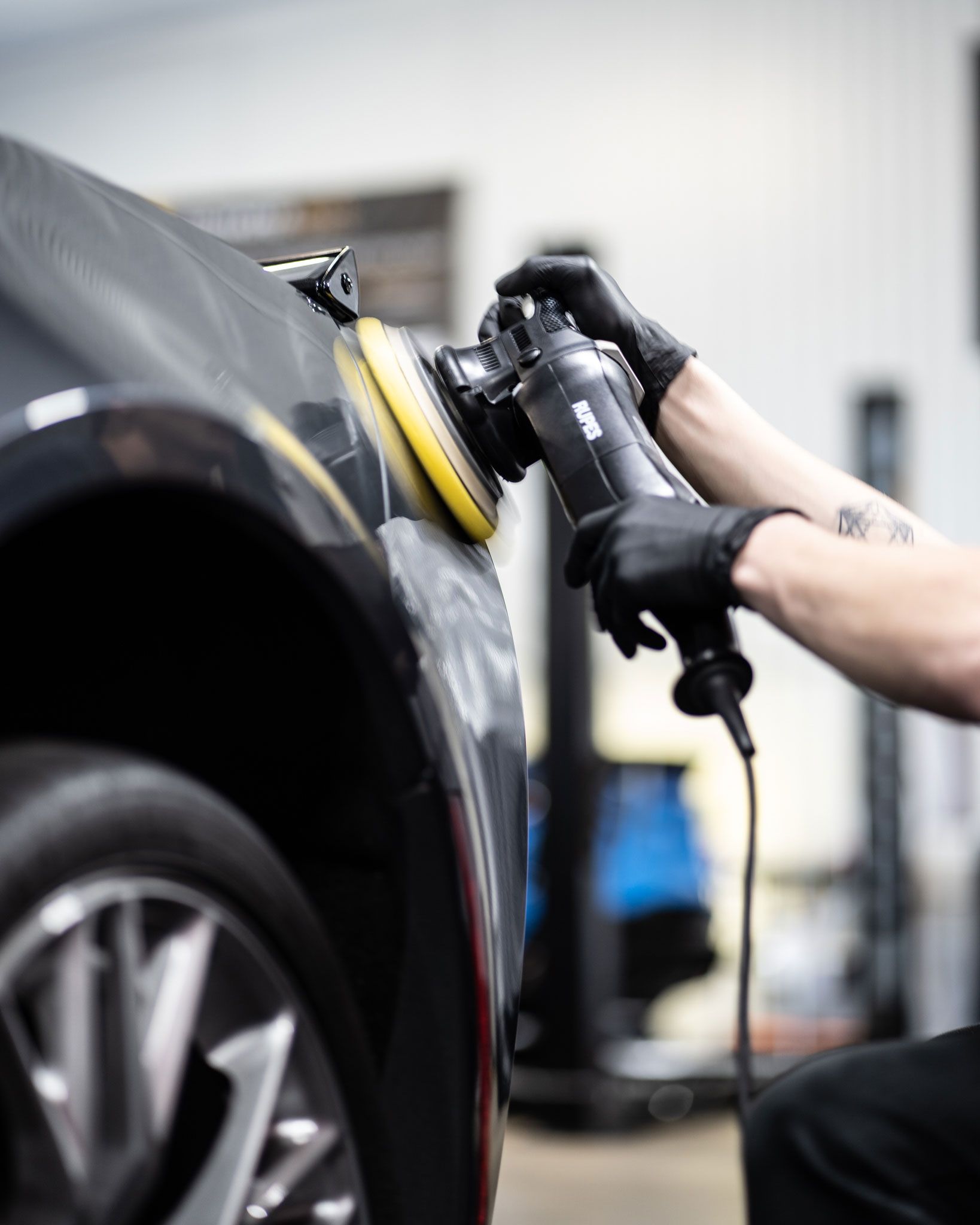 Person in black gloves polishing a black car with an electric buffer in a garage.