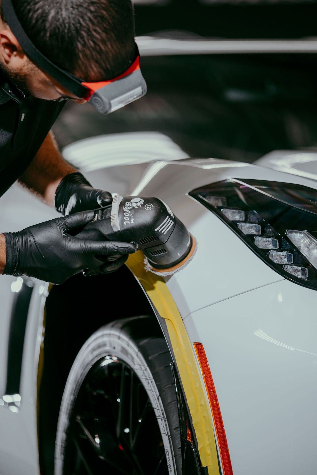 Man polishing a white sports car with a machine in a workshop, wearing black gloves and a headlamp.