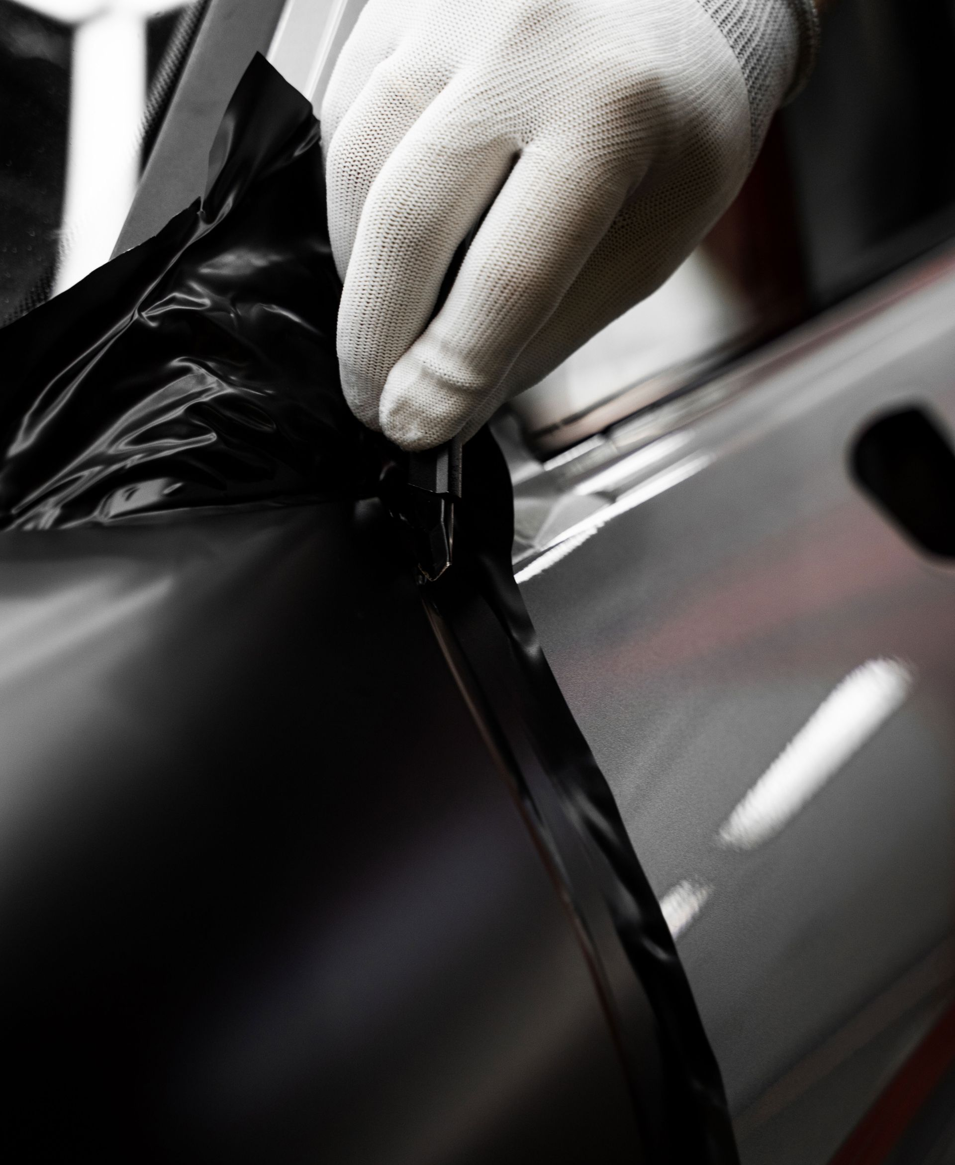 Gloved hand peeling black vinyl from a silver car panel, showing a car wrap installation.