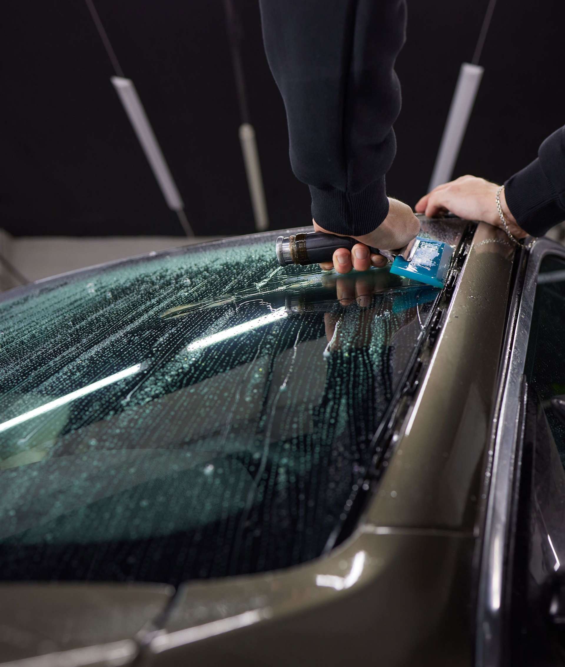 Person applying window tint to a car's windshield with a tool, inside a garage.