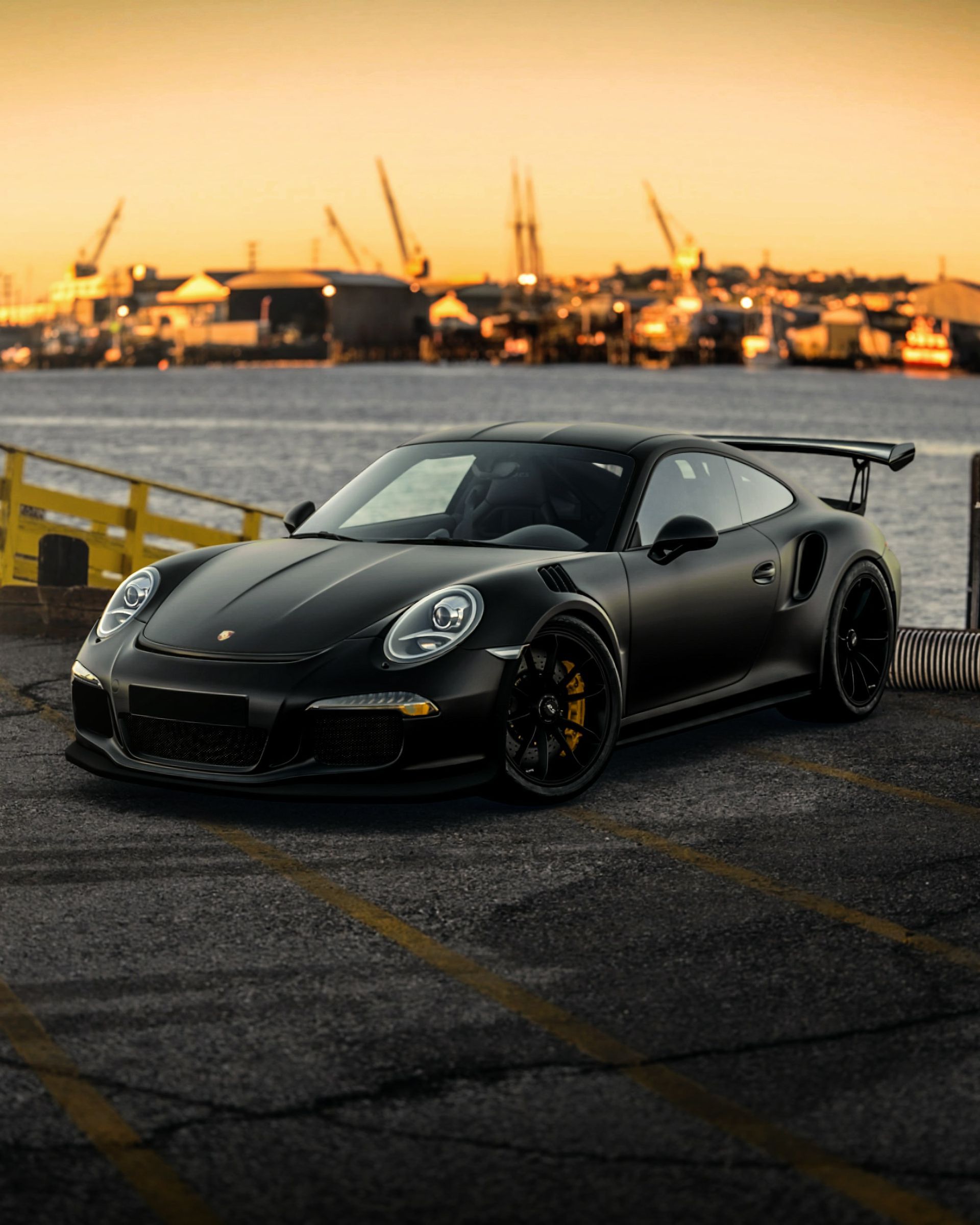 Black Porsche sports car parked at a harbor dock with a sunset in the background.
