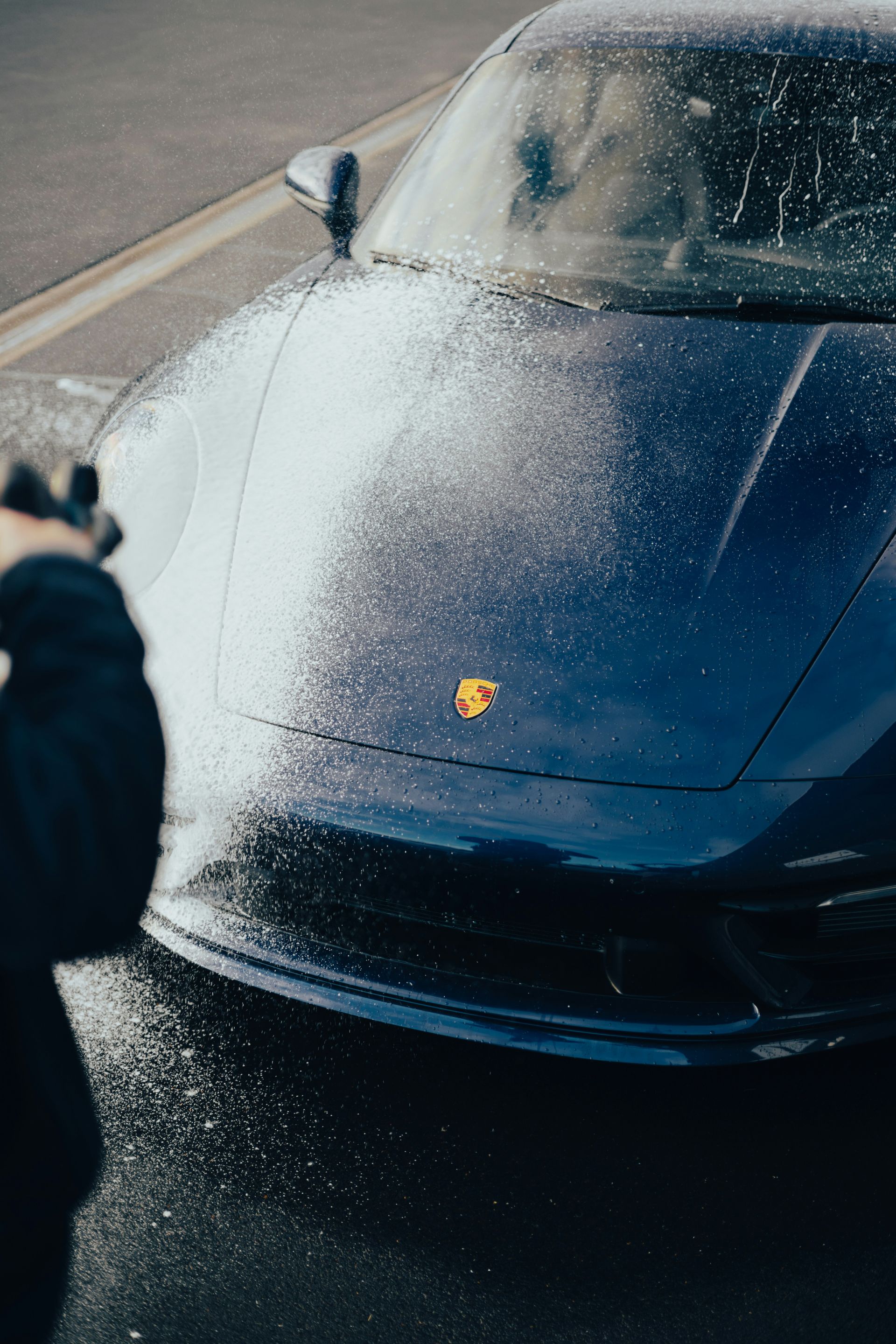 A blue Porsche being sprayed with water by a person in an outdoor setting.