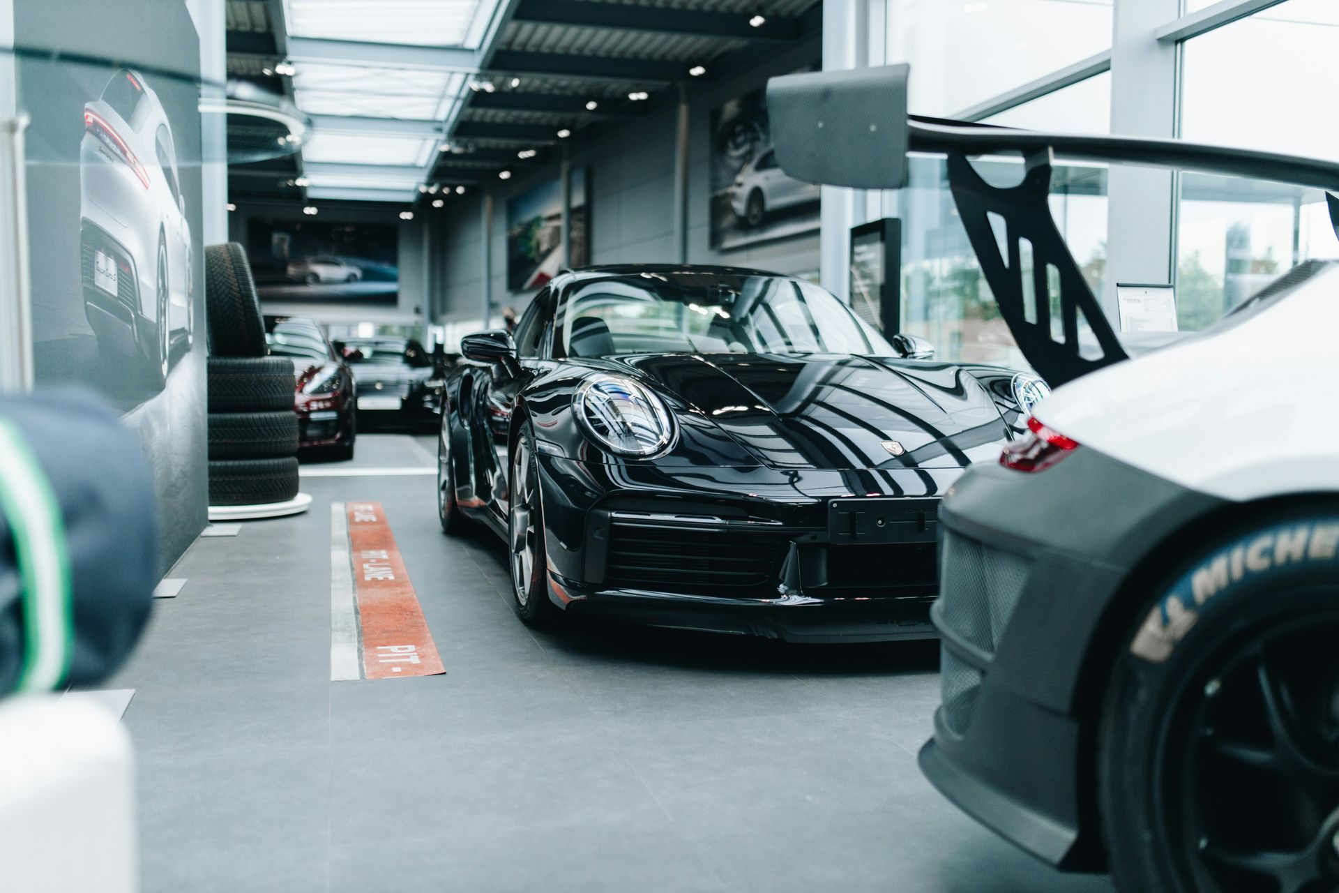 Black sports car in a showroom with other vehicles, tire, and a wing.