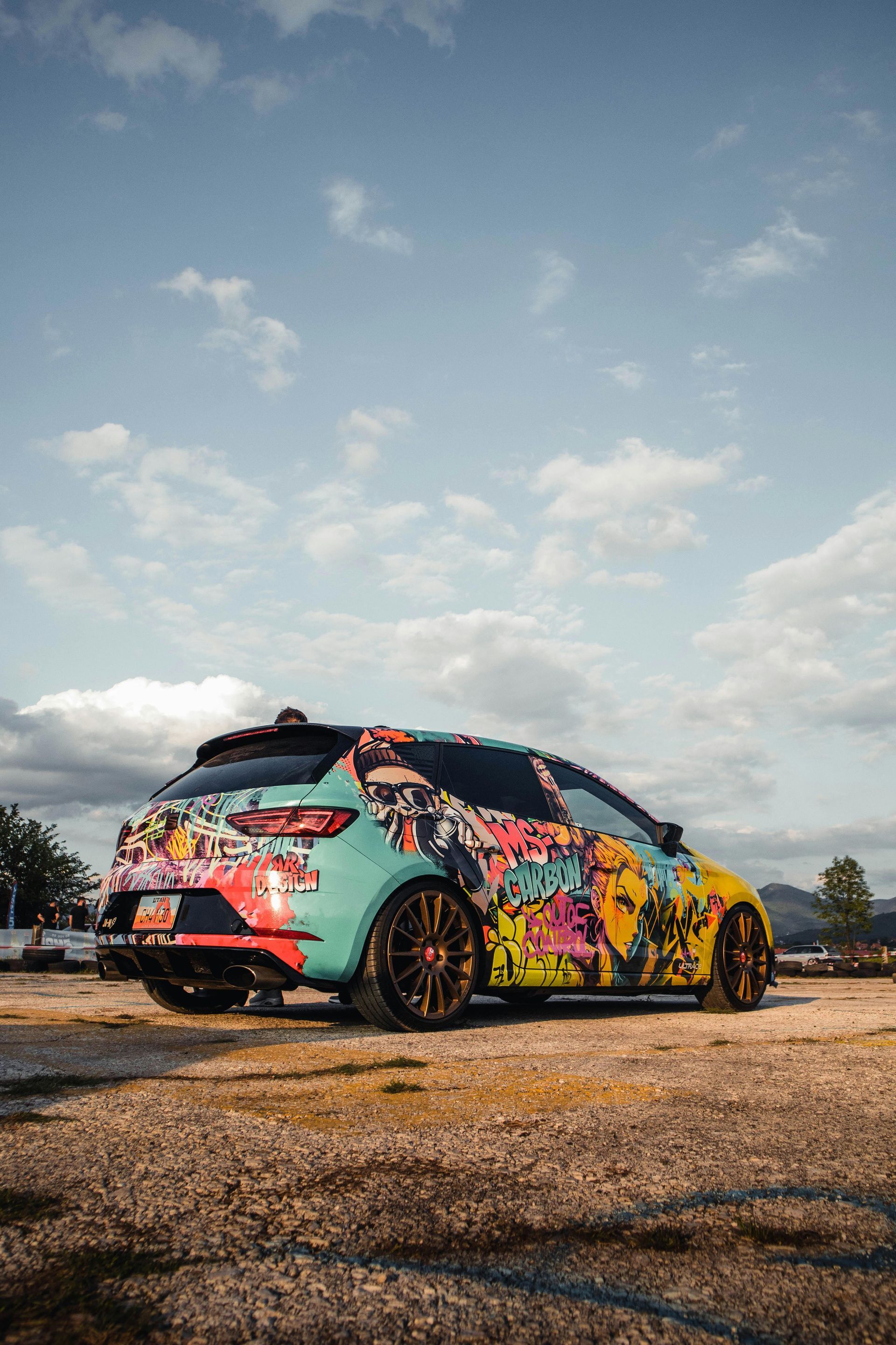 A vibrant, graffiti-style wrapped car parked on gravel under a blue sky.