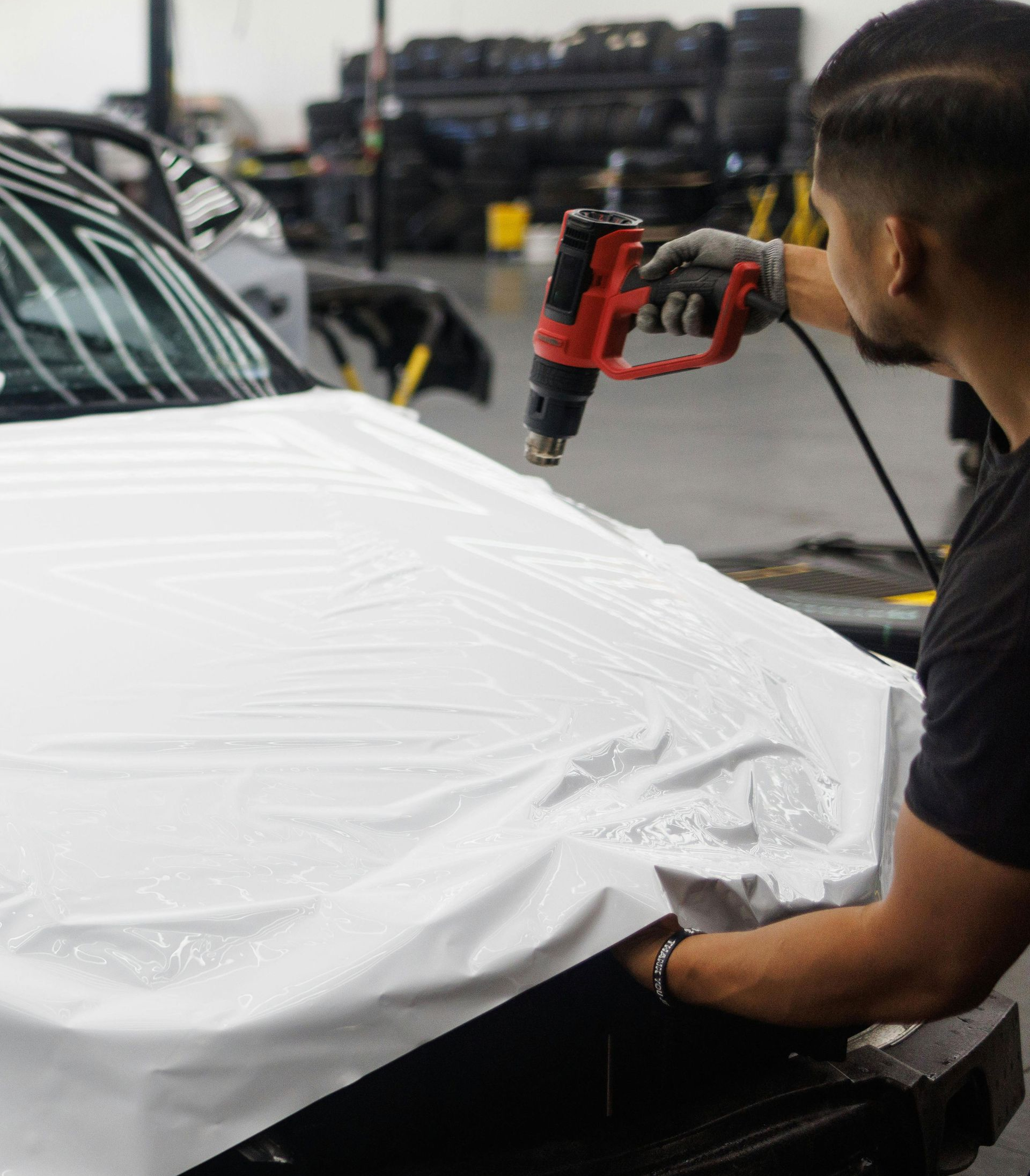 Man applying white vinyl wrap to a car hood with a heat gun in a garage.