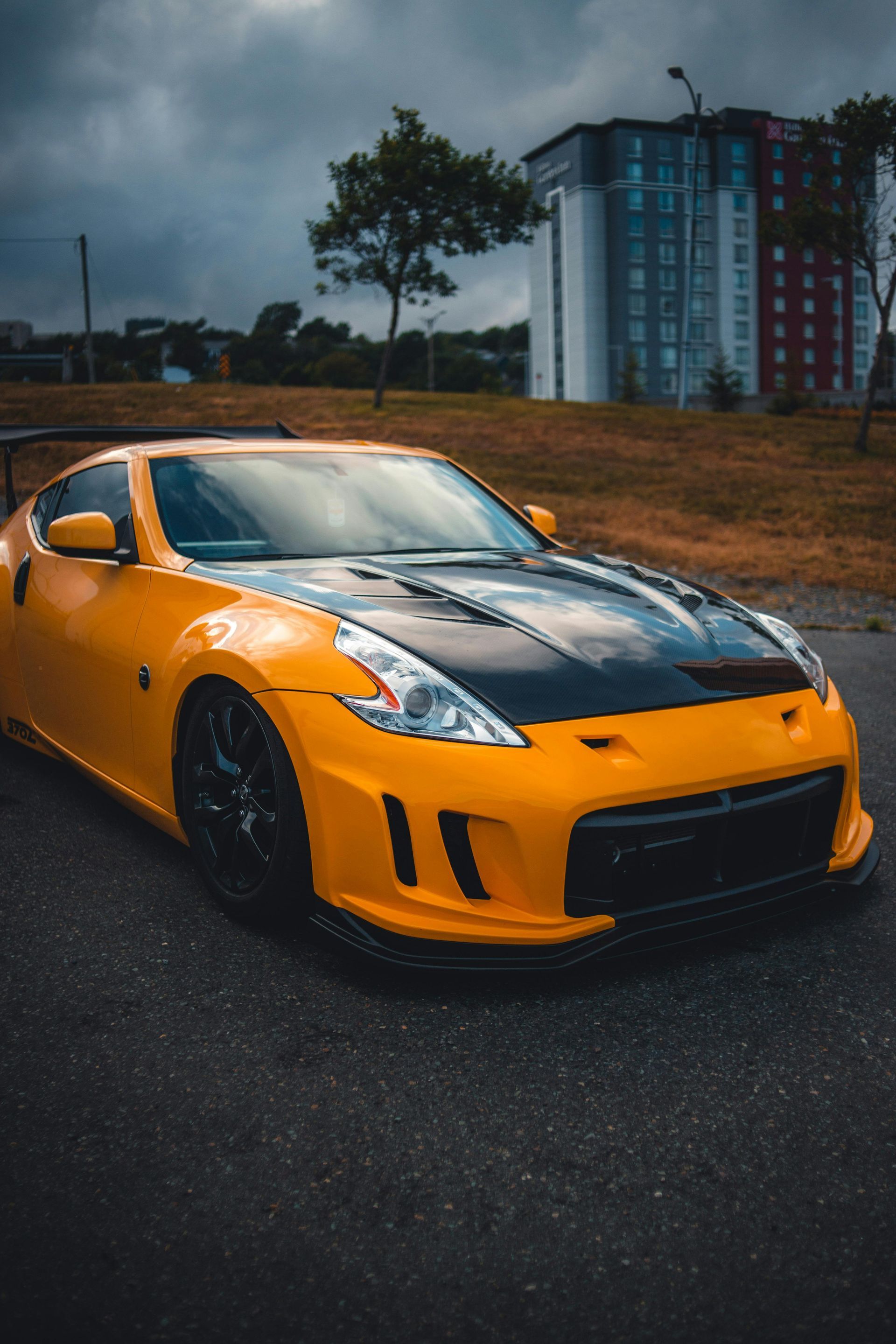 Yellow Nissan 370Z sports car with black hood, parked on asphalt in front of a building under a cloudy sky.