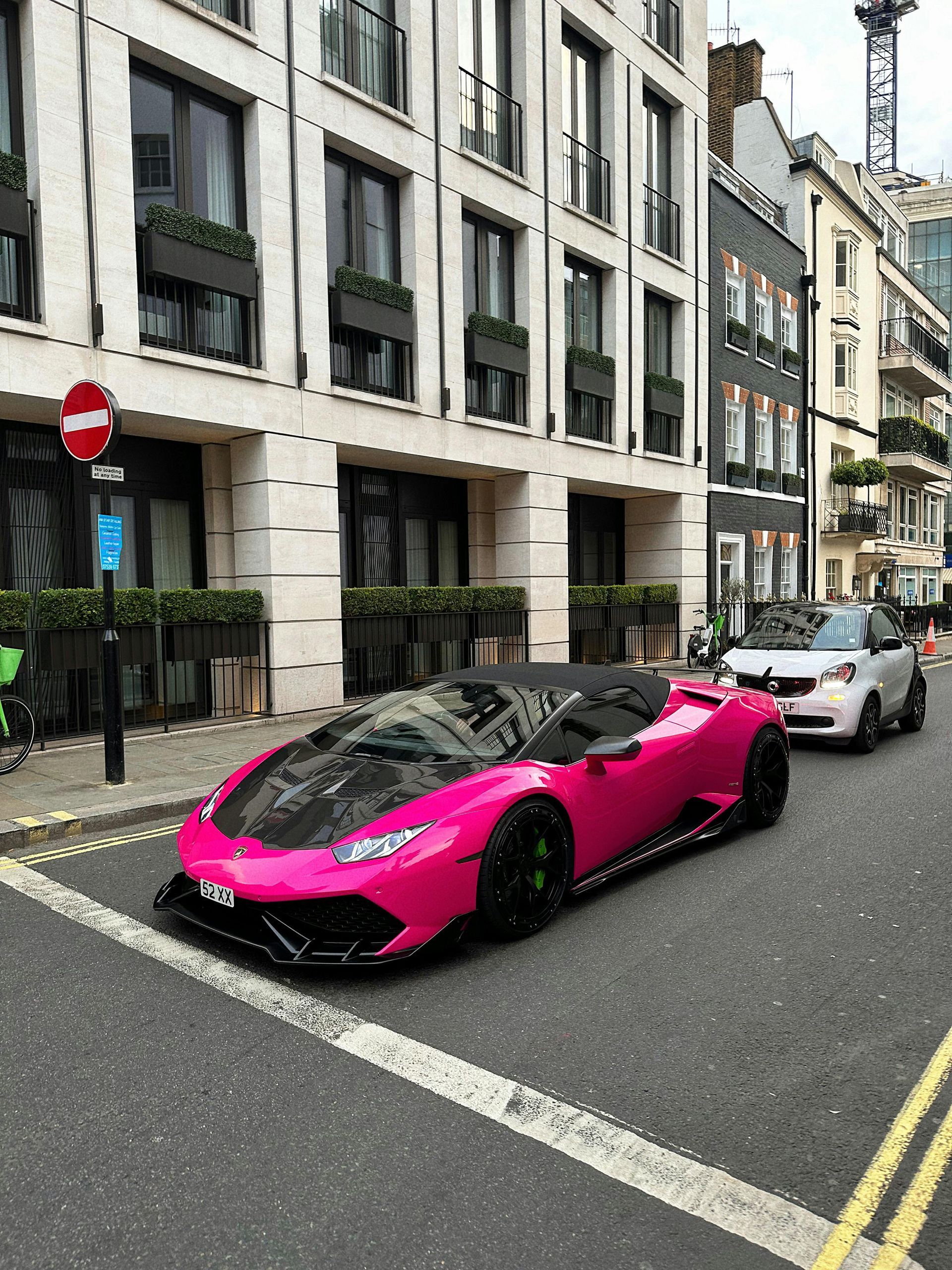 Pink Lamborghini convertible on a city street, black hood, with a white car behind it.