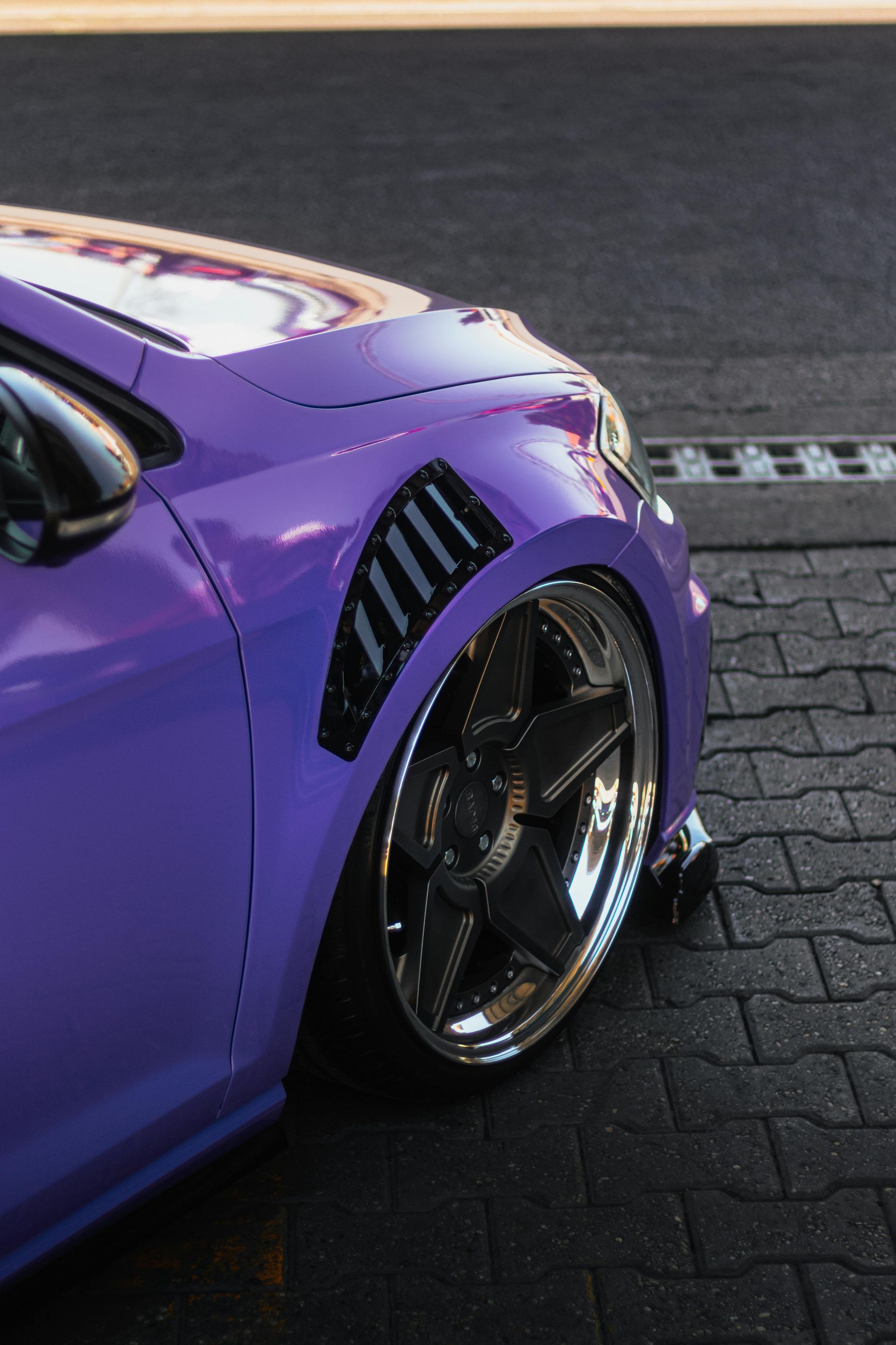 Purple car with custom fender vents and low profile chrome wheels, parked on a dark brick surface.