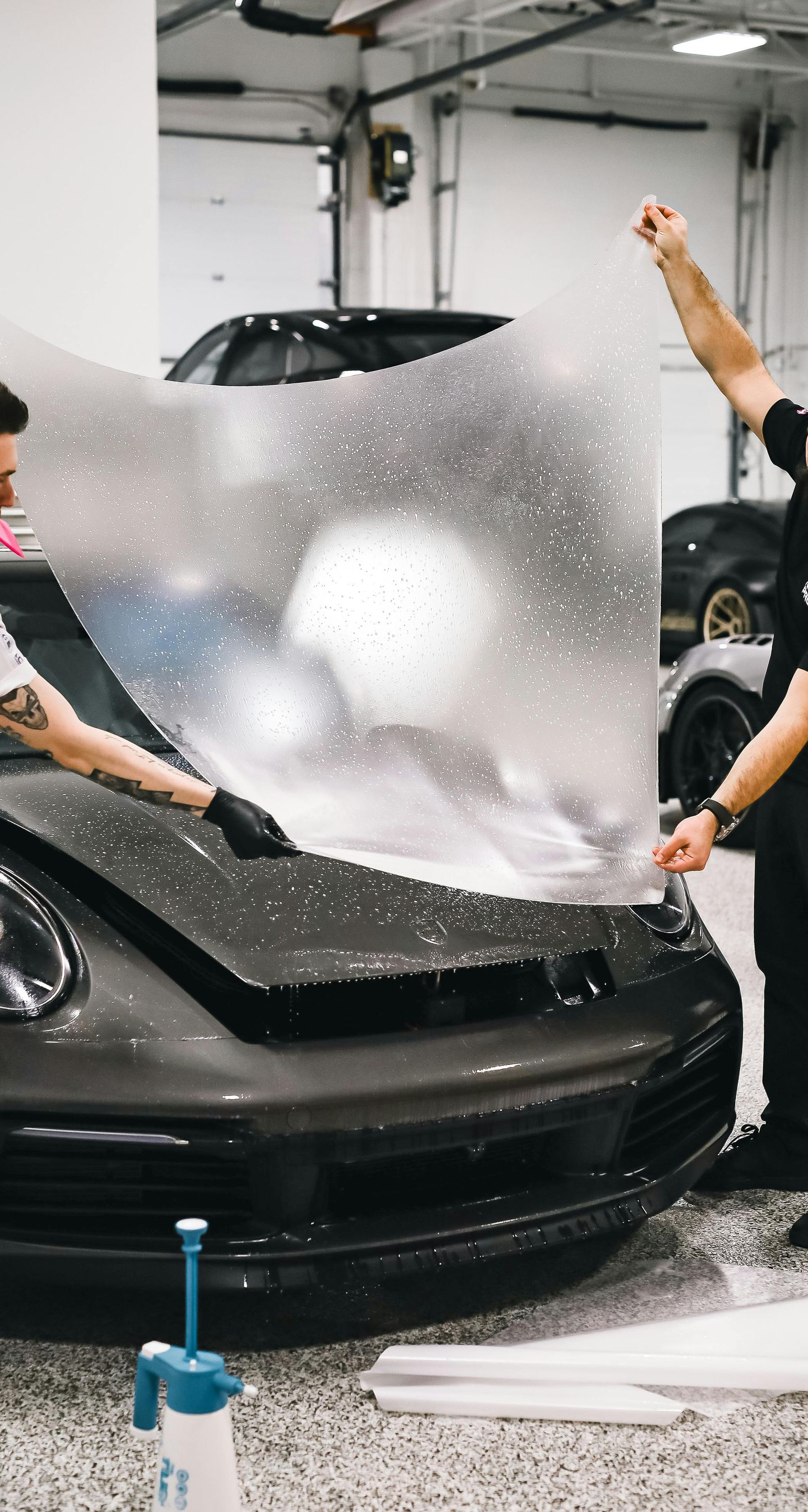 Two men applying protective film to a gray car hood in a garage.