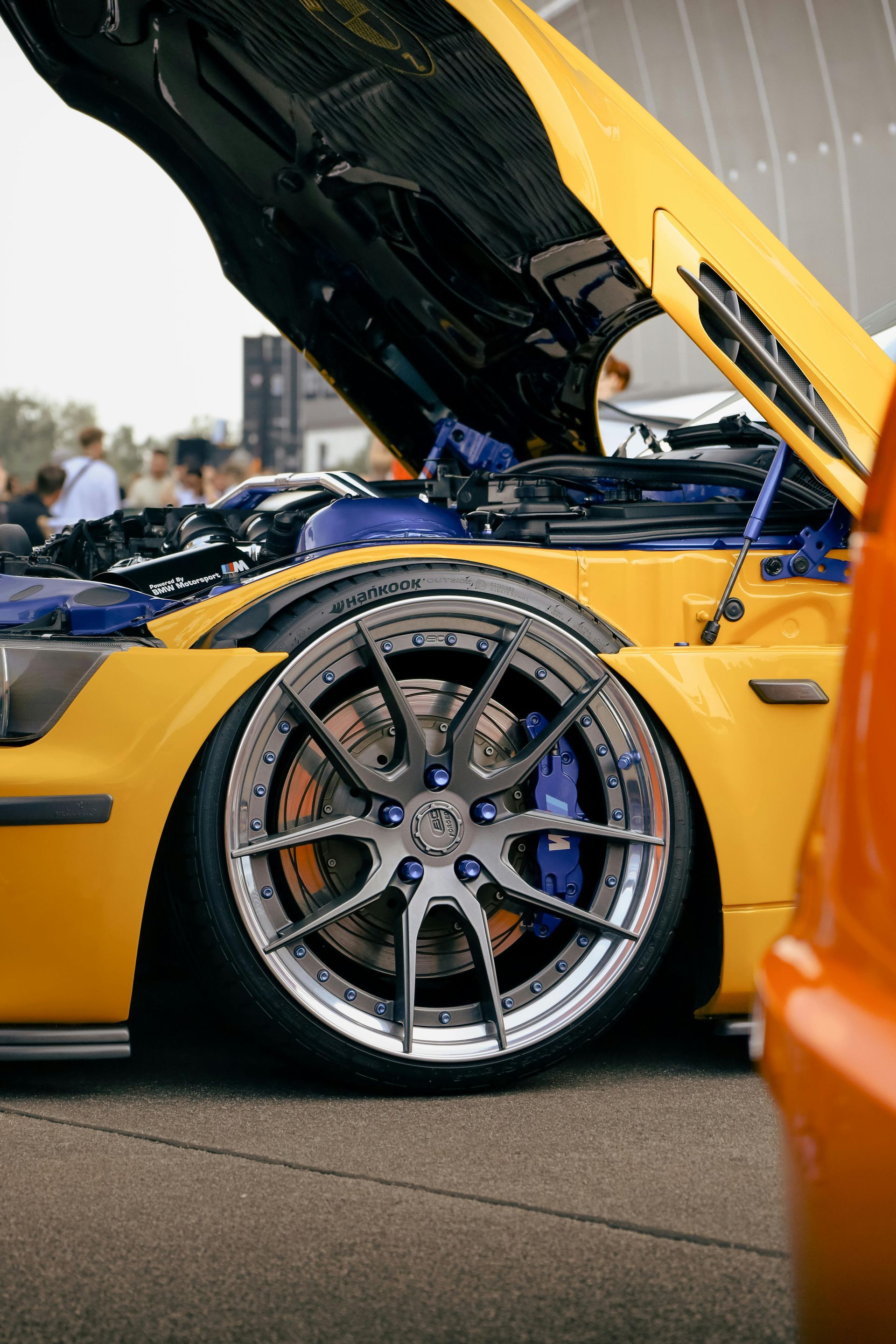 Yellow car with open hood, revealing engine. Chrome wheel, blue brakes.