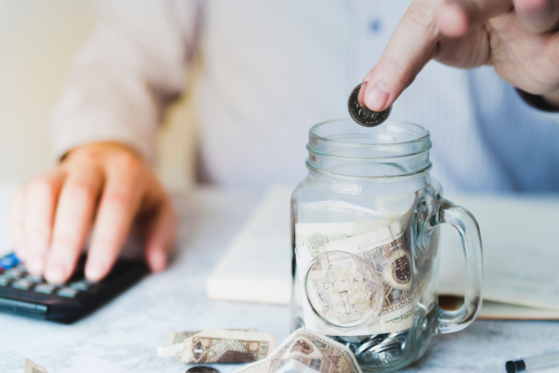 Une personne dépose une pièce dans un bocal en verre rempli d'argent, une calculatrice se trouve à proximité.