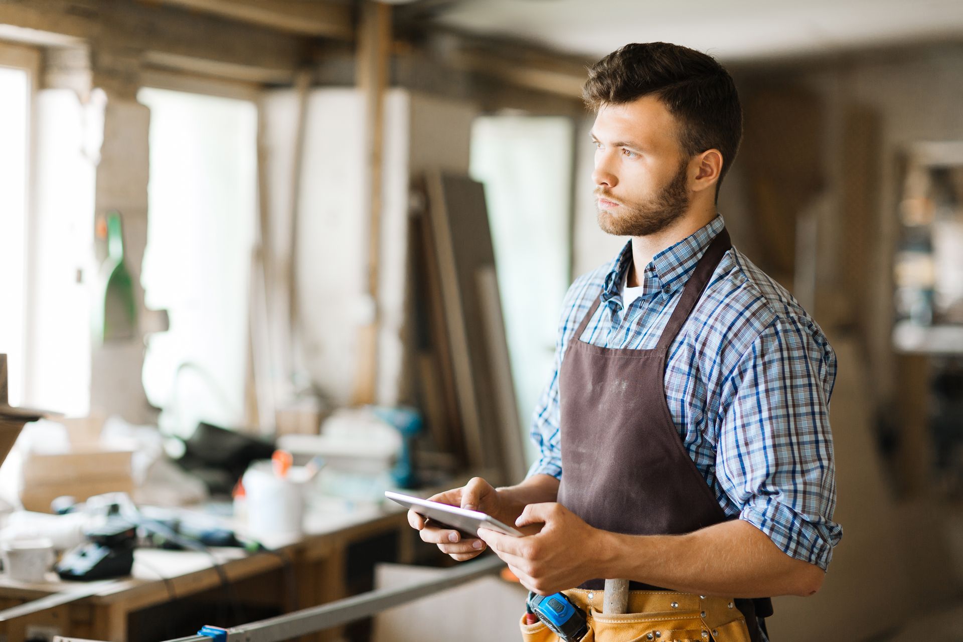 Menuisier dans son atelier, l'air pensif, tenant une tablette, portant un tablier, établi en arrière-plan.