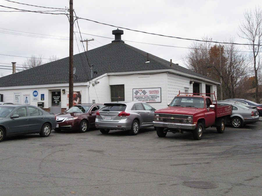 Cars parked outside a white auto shop with a dark roof and red pickup truck in front | Village Repair